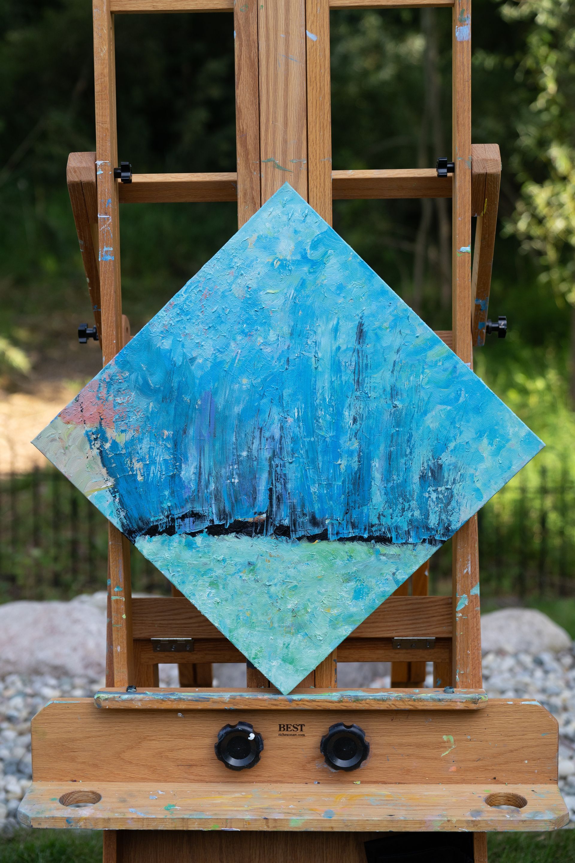 Abstract blue and green painting on a diamond-shaped canvas, mounted on a wooden easel outdoors.