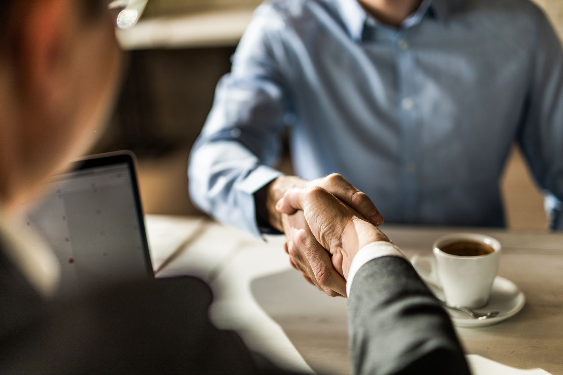 Two men are shaking hands over a table with a cup of coffee .