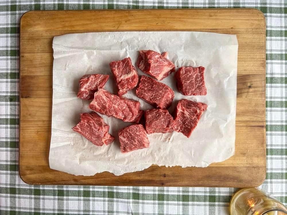 A wooden cutting board topped with pieces of raw beef on a checkered table cloth.