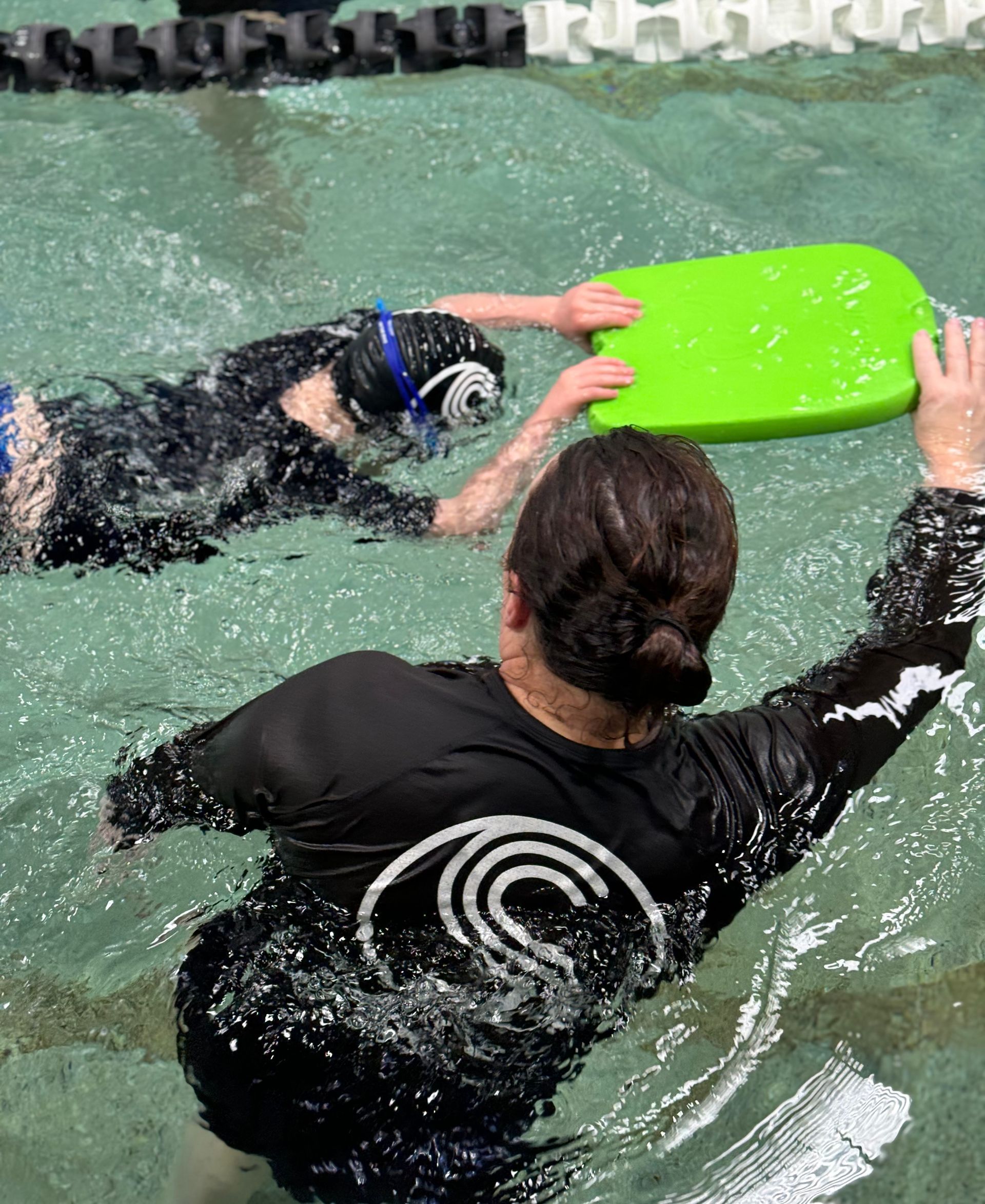 A woman in a black shirt is holding a green board in a swimming pool