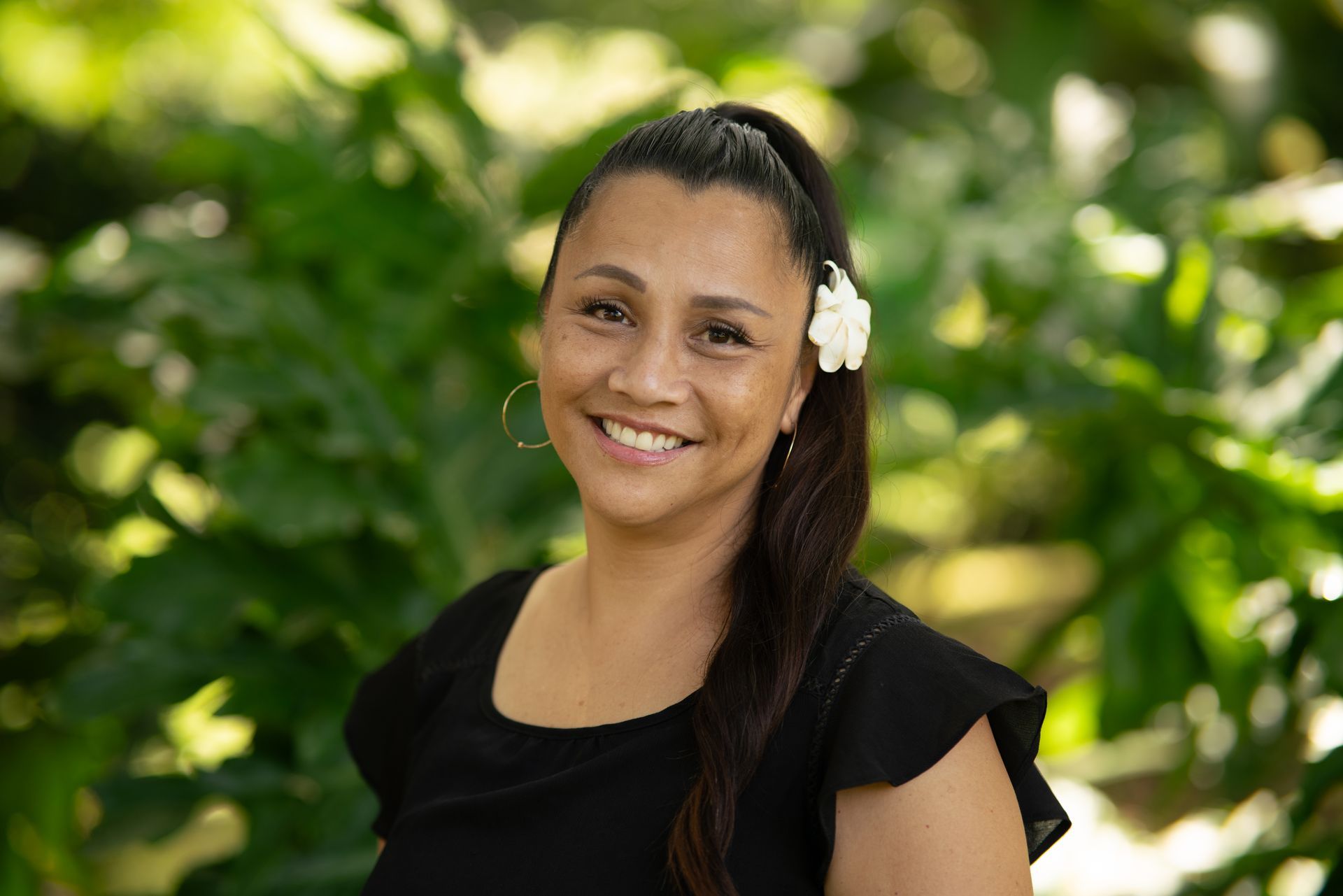 Smiling woman in a black top with a white flower in her hair, standing outdoors with greenery behind her Smiling woman in a black top with a white flower in her hair, standing outdoors with greenery behind her