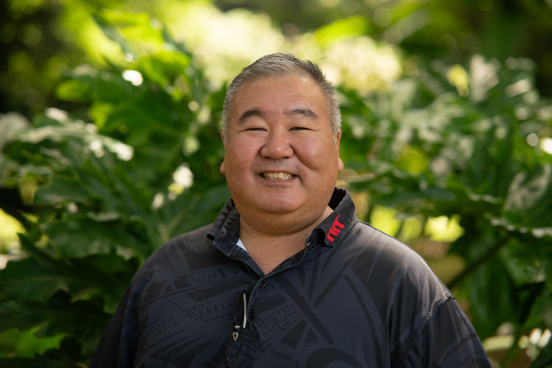 Smiling person in a dark jacket standing outdoors with green foliage behind them Smiling person in a dark jacket standing outdoors with green foliage behind them