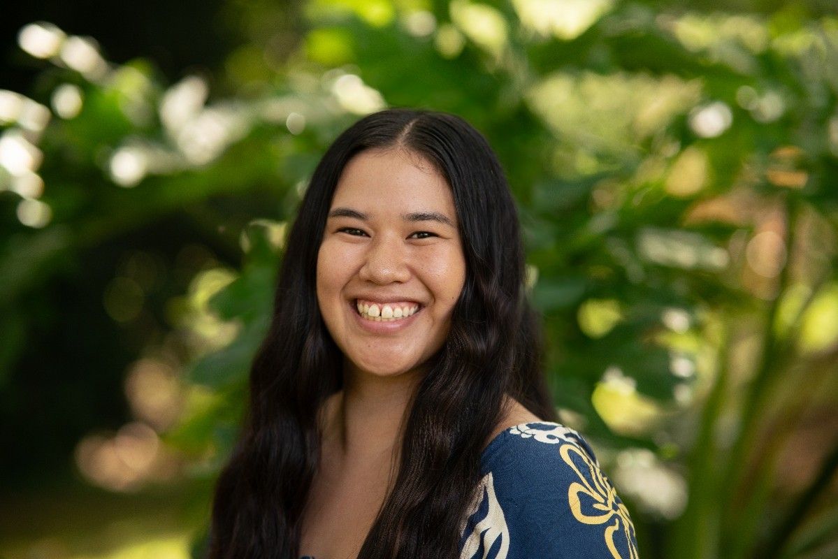 Smiling woman outdoors with long dark hair and a blue patterned top, with green foliage behind her Smiling woman outdoors with long dark hair and a blue patterned top, with green foliage behind her