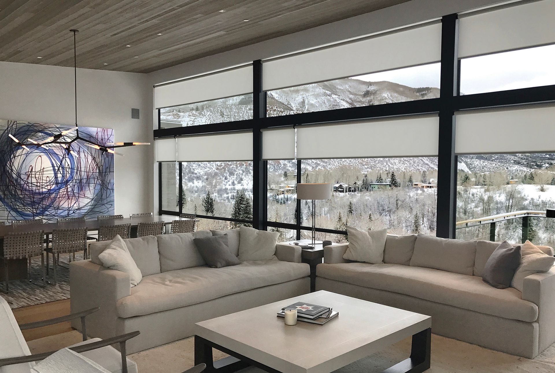 Living room with snow-covered mountain view; neutral-toned sofas, large painting, and window shades.