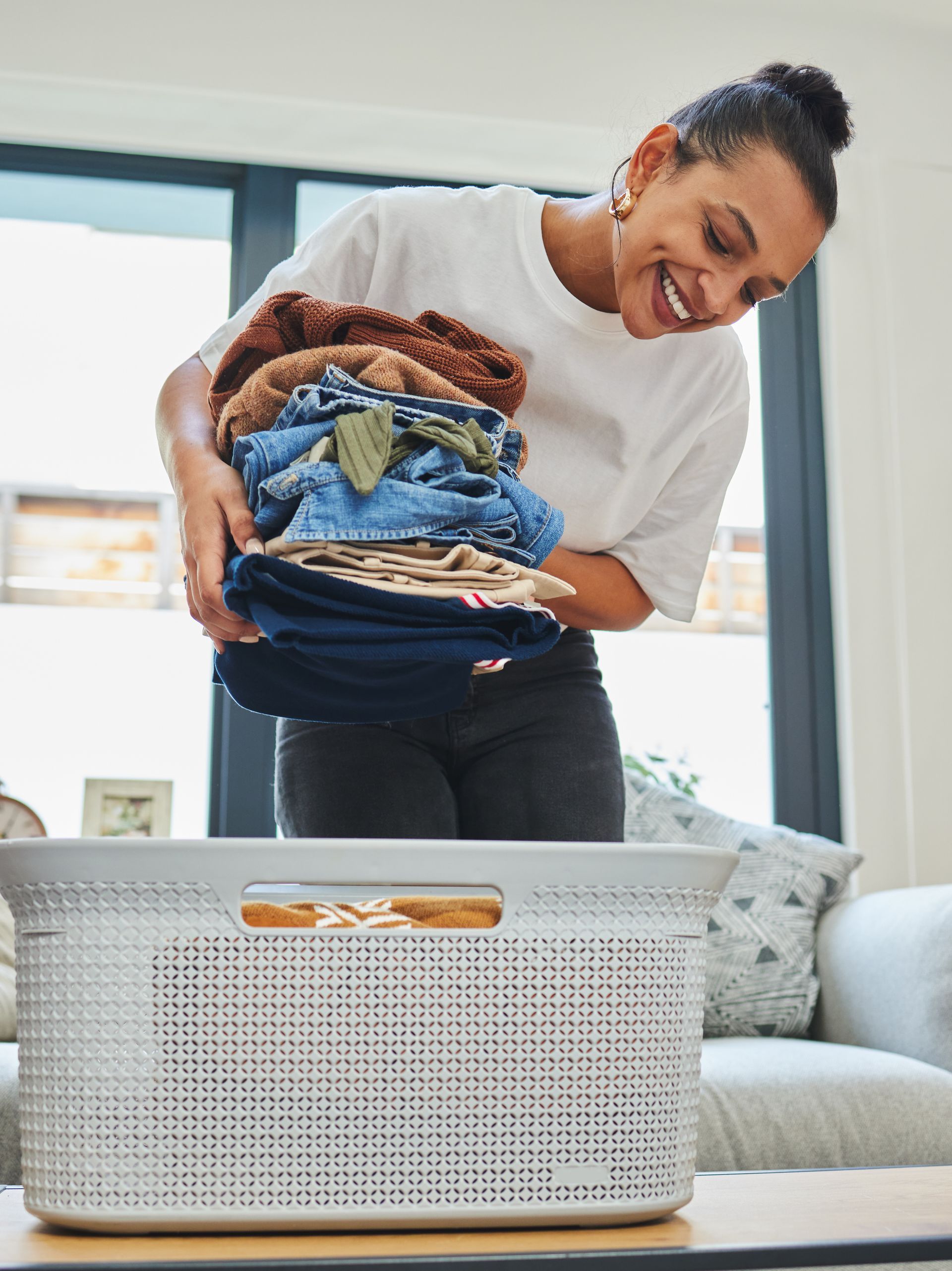 Woman placing folded laundry into a white basket in a bright living room