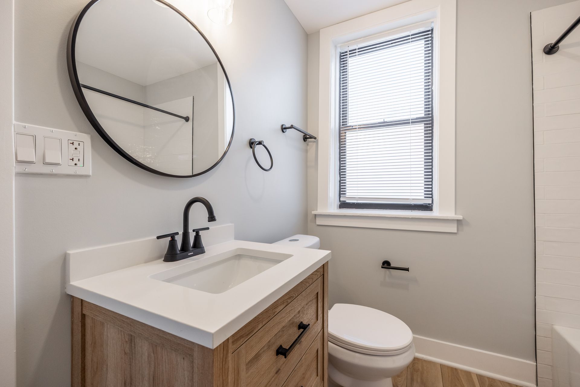 Small modern bathroom with round mirror, wood vanity, white sink, toilet, and window.