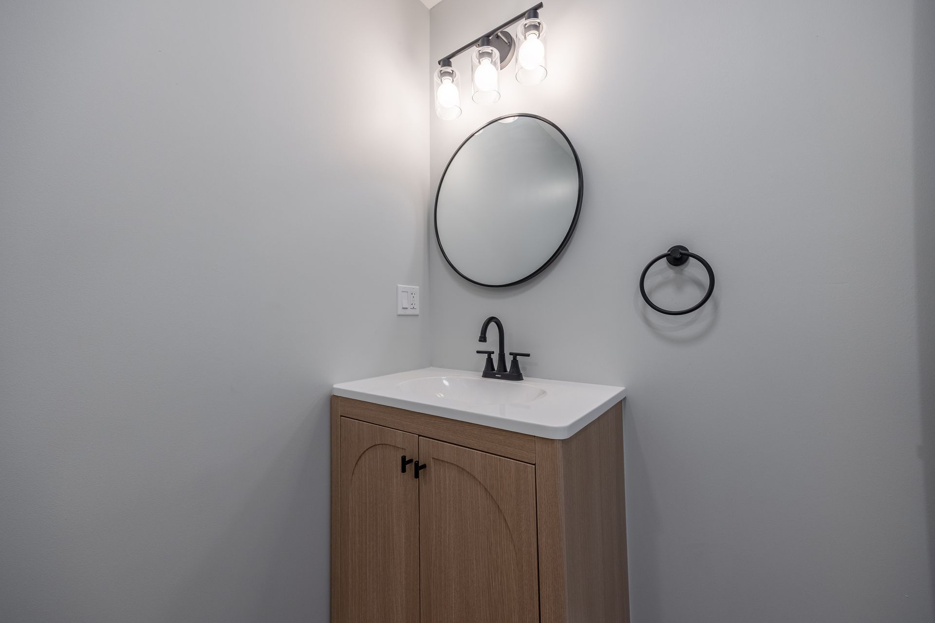 Small bathroom vanity with round mirror, black faucet, and towel ring on light gray walls.