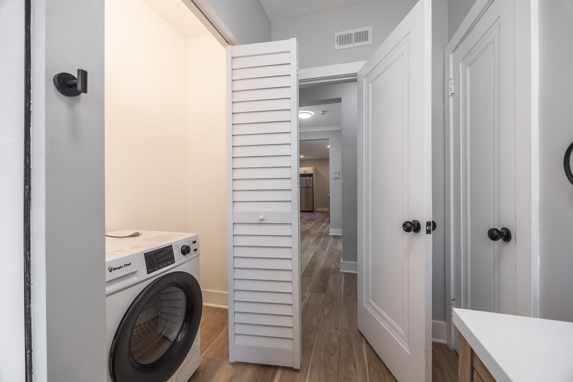 Laundry room hallway with a front-loading washer and white louvered doors in a narrow white space
