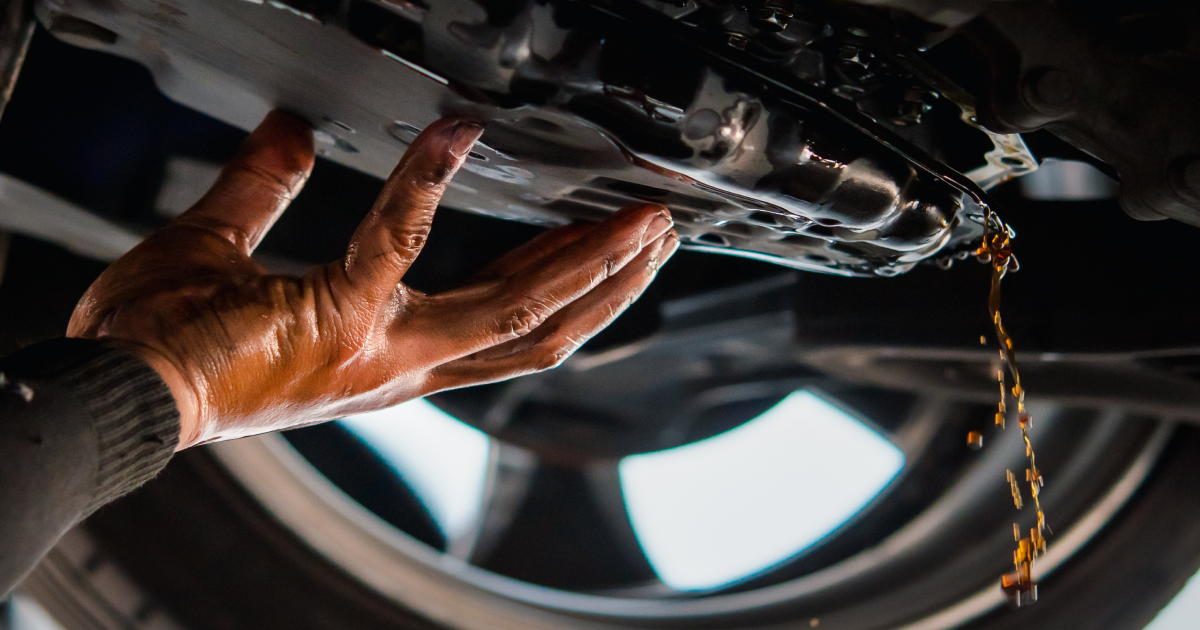 A person's oil-stained hand adjusting a metal plate under a vehicle as dark fluid drips from the car.