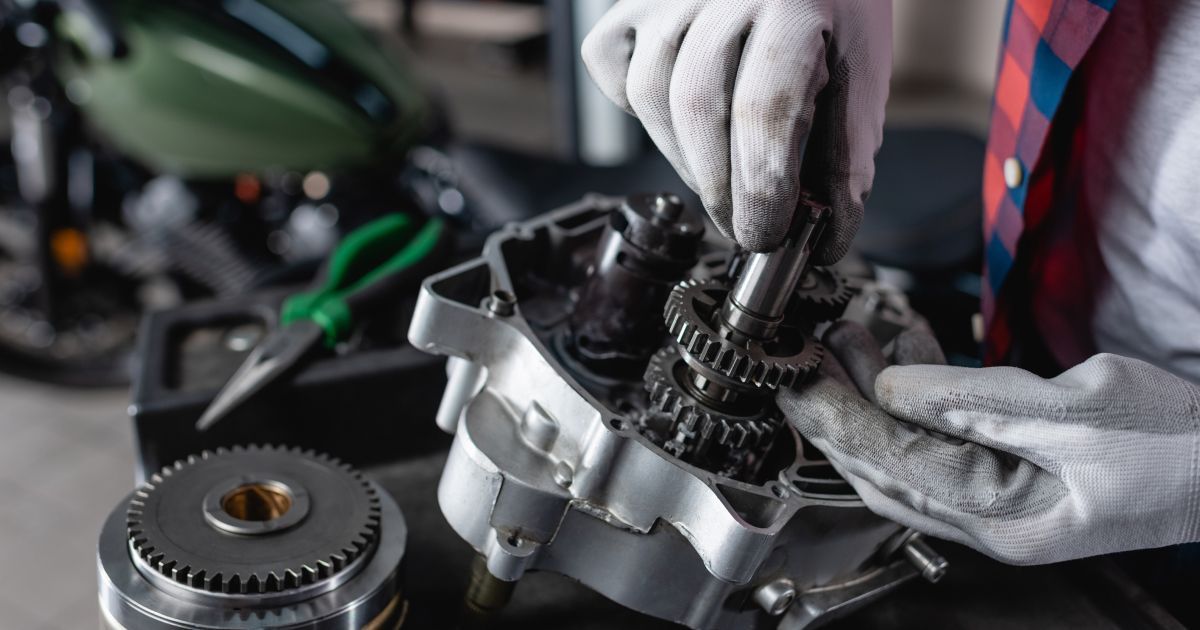 A gloved hand assembles a metallic gear mechanism inside a motorcycle engine block in a repair shop.