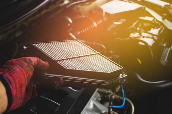 A mechanic wearing a red plaid glove holds a used, rectangular car air filter over an open engine bay.