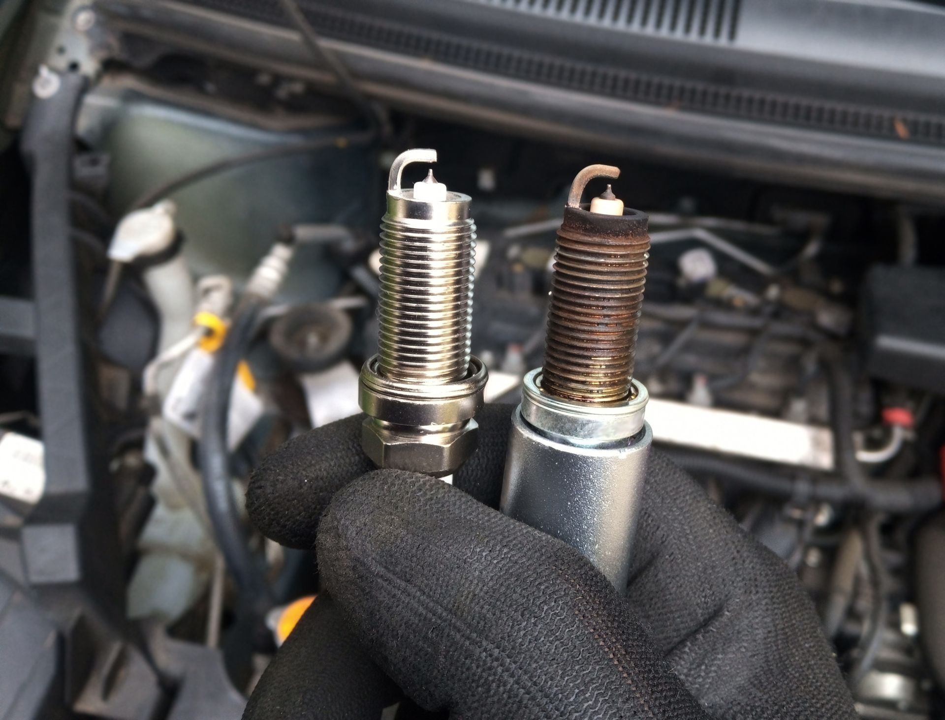 A gloved hand holds a new, clean spark plug next to a used, worn one in front of an open car engine bay.
