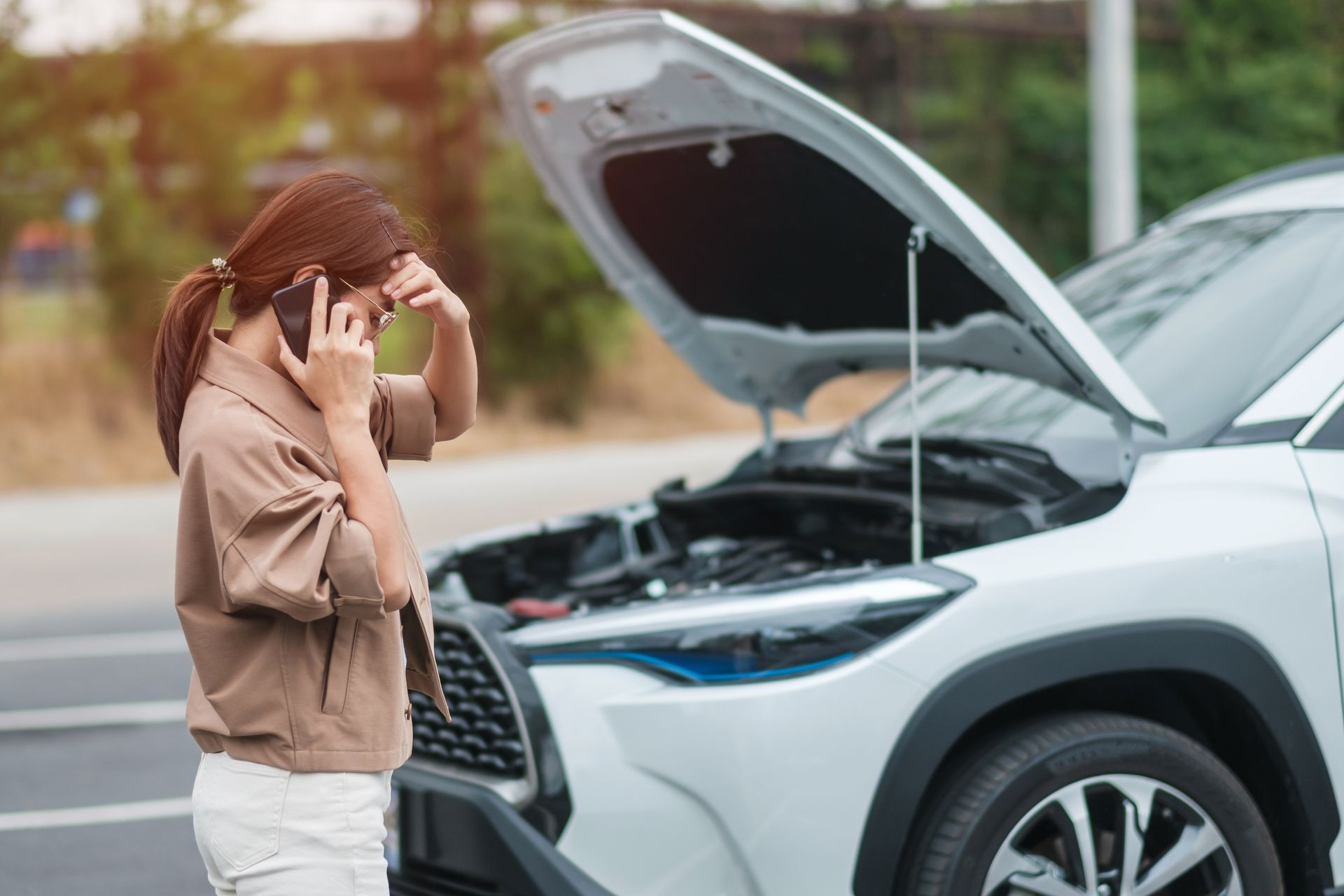 A person standing on the side of a road on a mobile phone looking stressed in front of a car with its hood open.