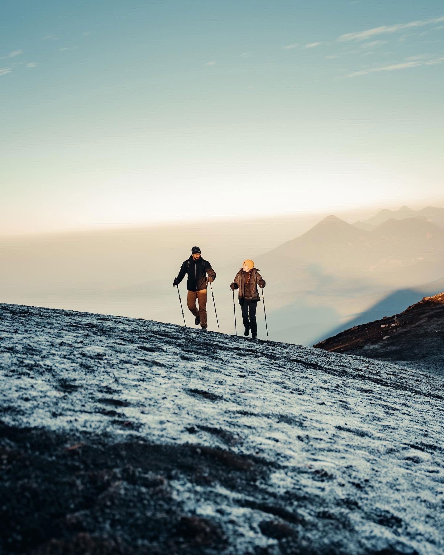 Two hikers with trekking poles climb a snowy mountain slope at sunrise.