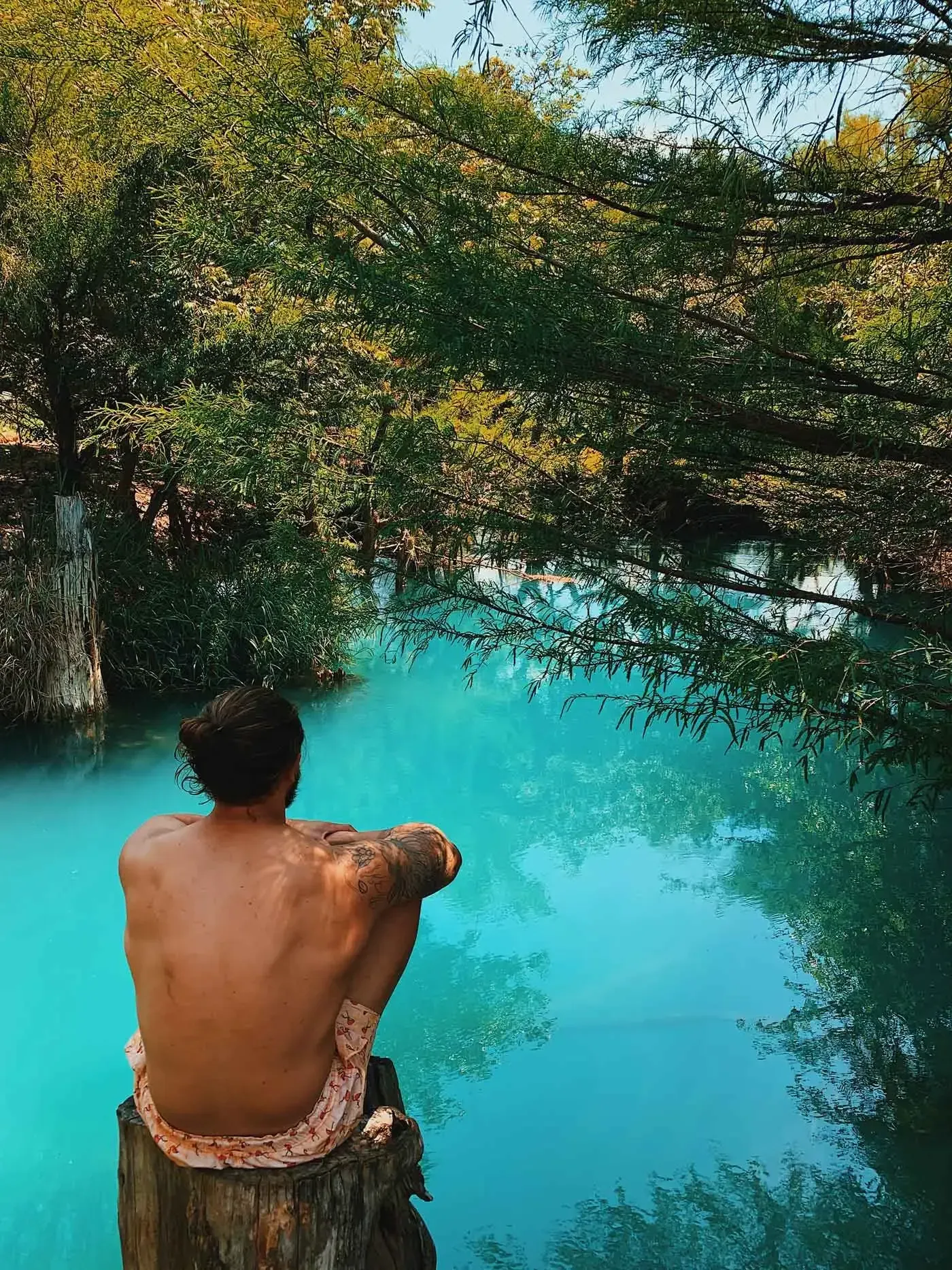 Person sits on a rock overlooking a bright turquoise river surrounded by dense trees.