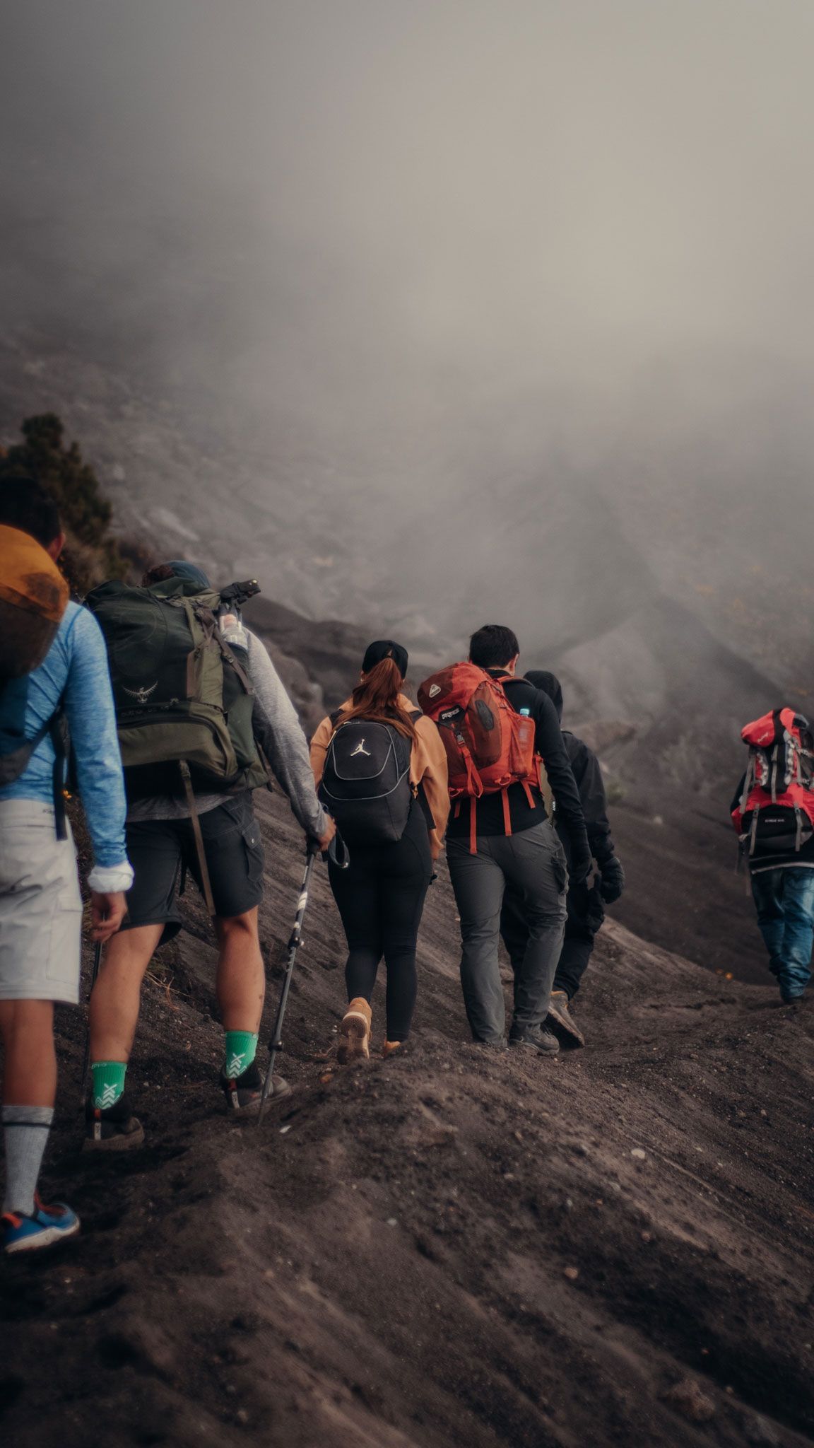 Hikers with backpacks walking along a muddy volcanic trail in dense fog