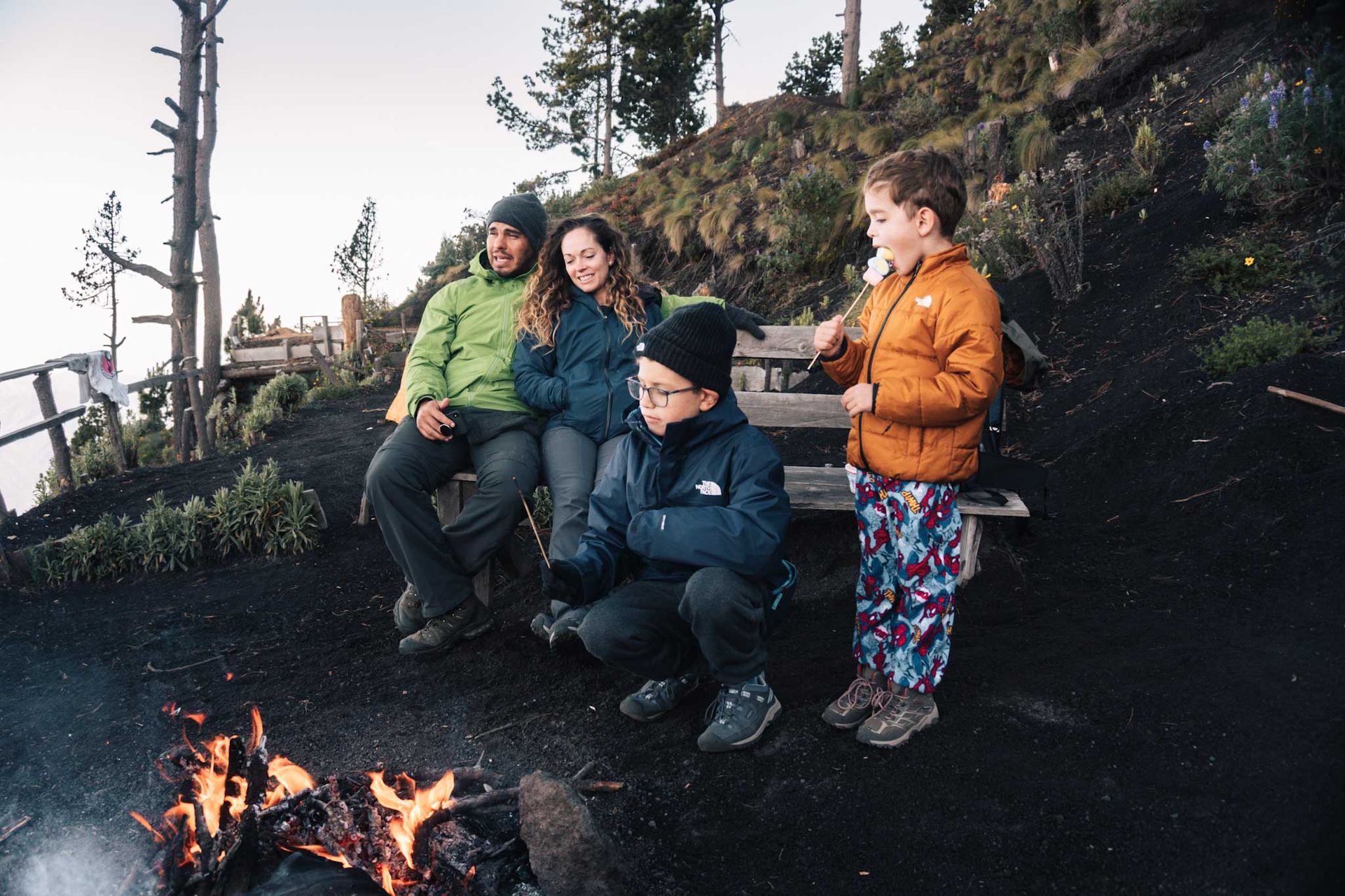 Four people sit and stand by a small campfire outdoors on a rocky hillside at dusk.