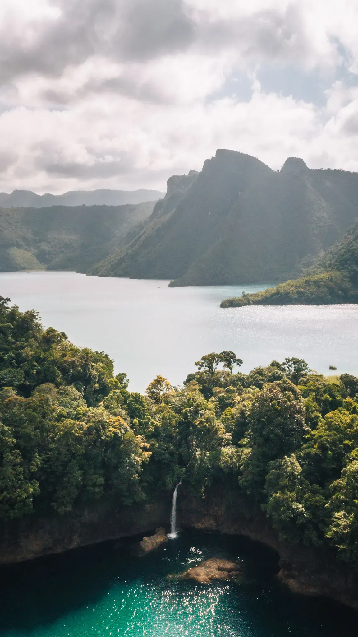 Lush tropical cove with turquoise water, dense green forest, and misty mountains under cloudy sky