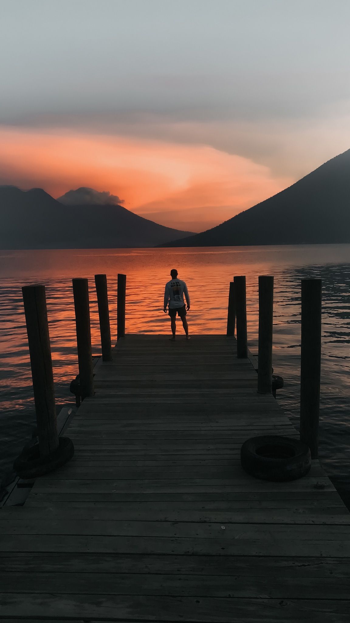 Person standing on a wooden dock at sunset with mountains and orange sky reflected on the water