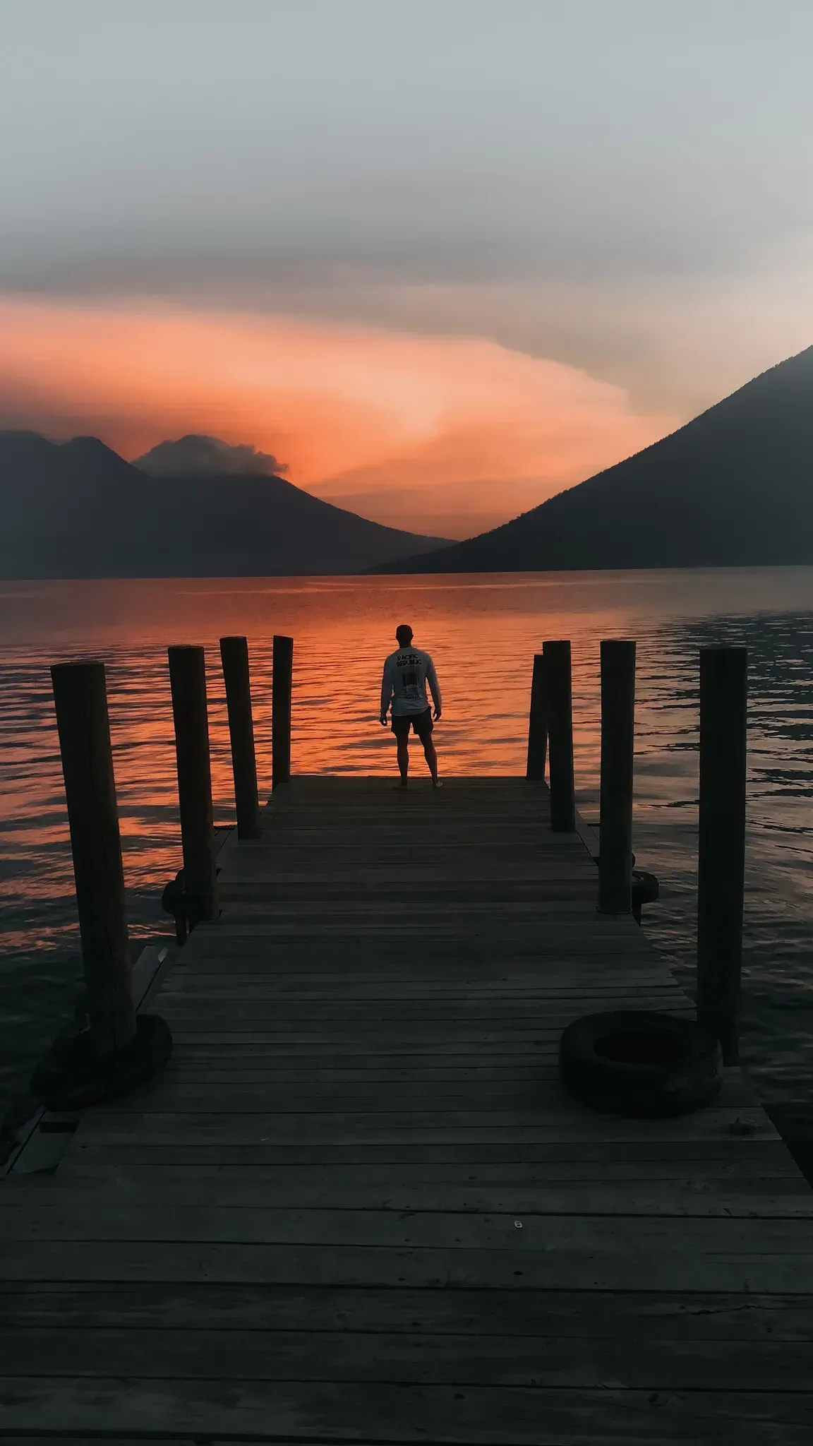Person standing on a wooden dock at sunset, with orange sky and mountains across the water.