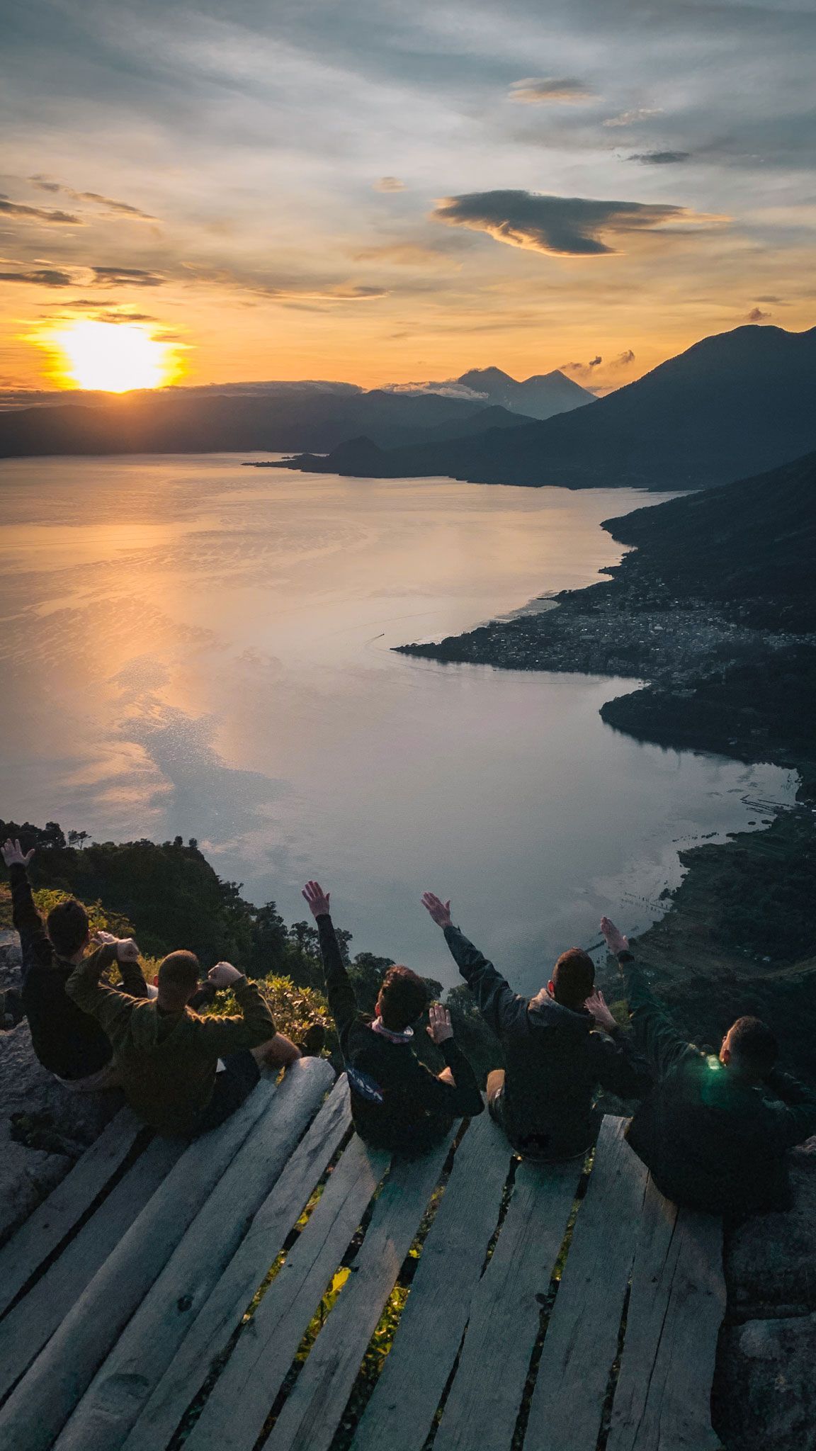 Sunset over a mountain lake, with silhouettes of people seated on a hill overlooking the water.