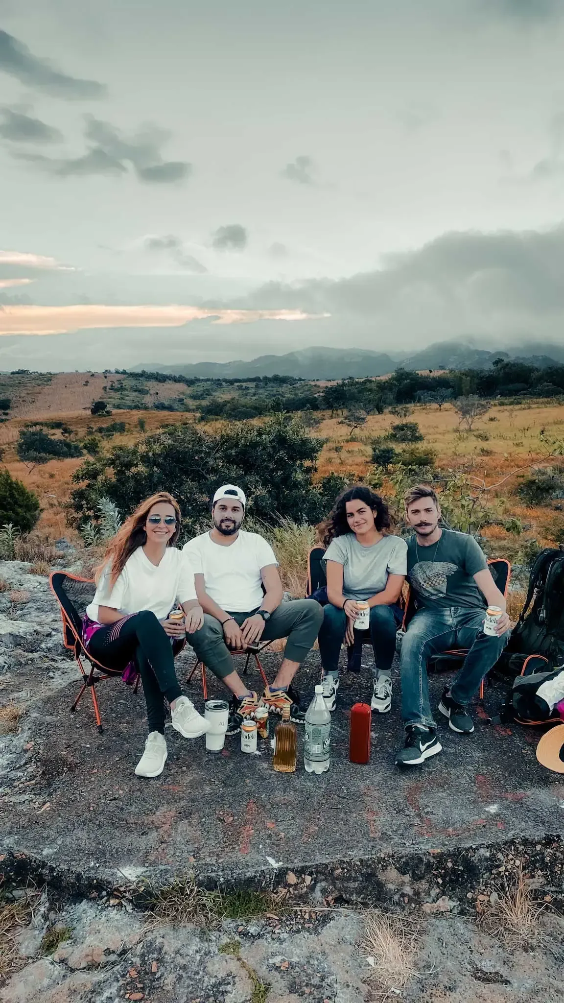 Five people sitting outdoors on a rocky ledge at dusk, with drinks and a scenic landscape behind them