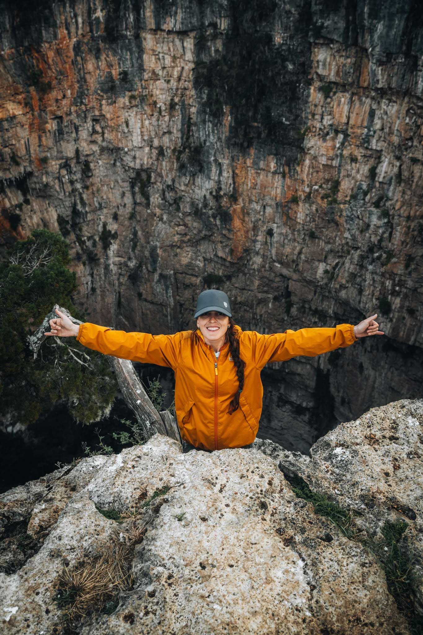 Person in an orange jacket and cap stands with arms outstretched among rocky cliffs.