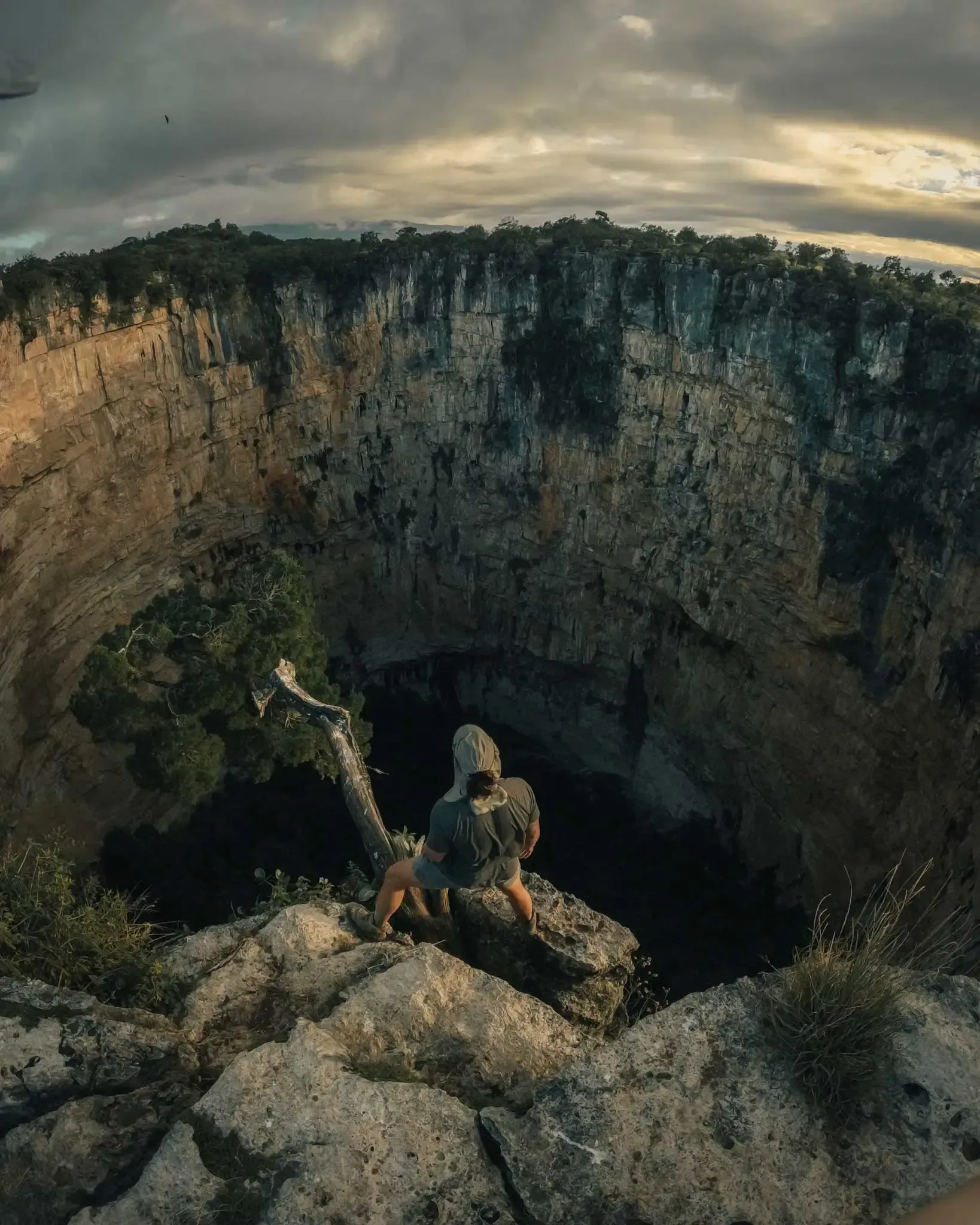 Two people sit on a rocky cliff edge above a deep volcanic crater under a cloudy sky.