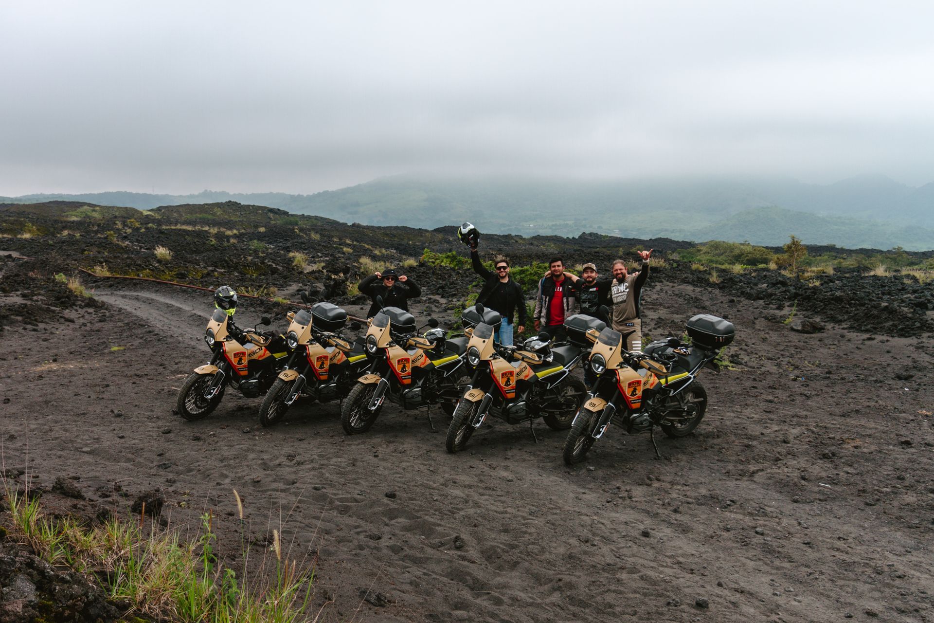 Four off-road motorcycles parked on a rocky trail with riders in the background under a cloudy sky