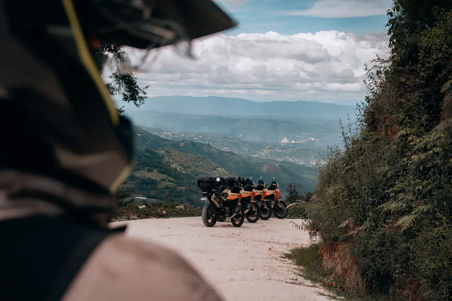 Three motorcycles parked on a mountain road overlooking a valley under a cloudy sky