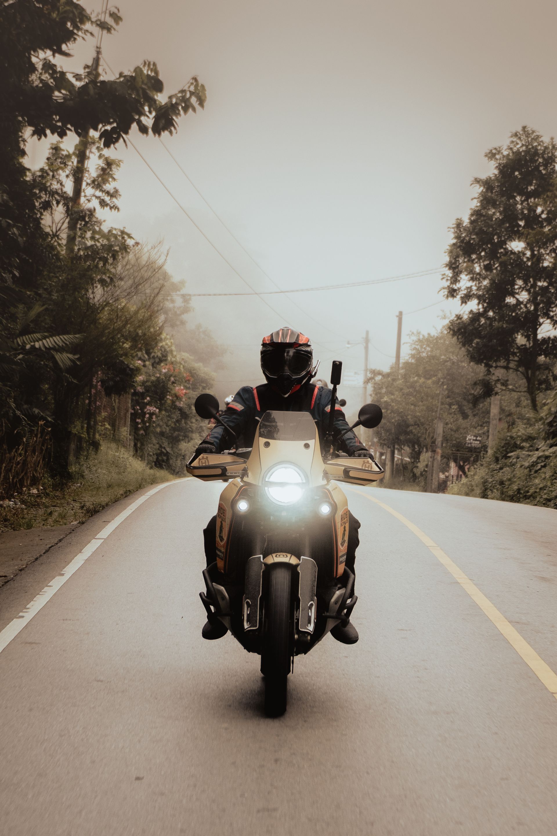 Motorcyclist riding toward camera on a tree-lined road in hazy daylight, headlight on.