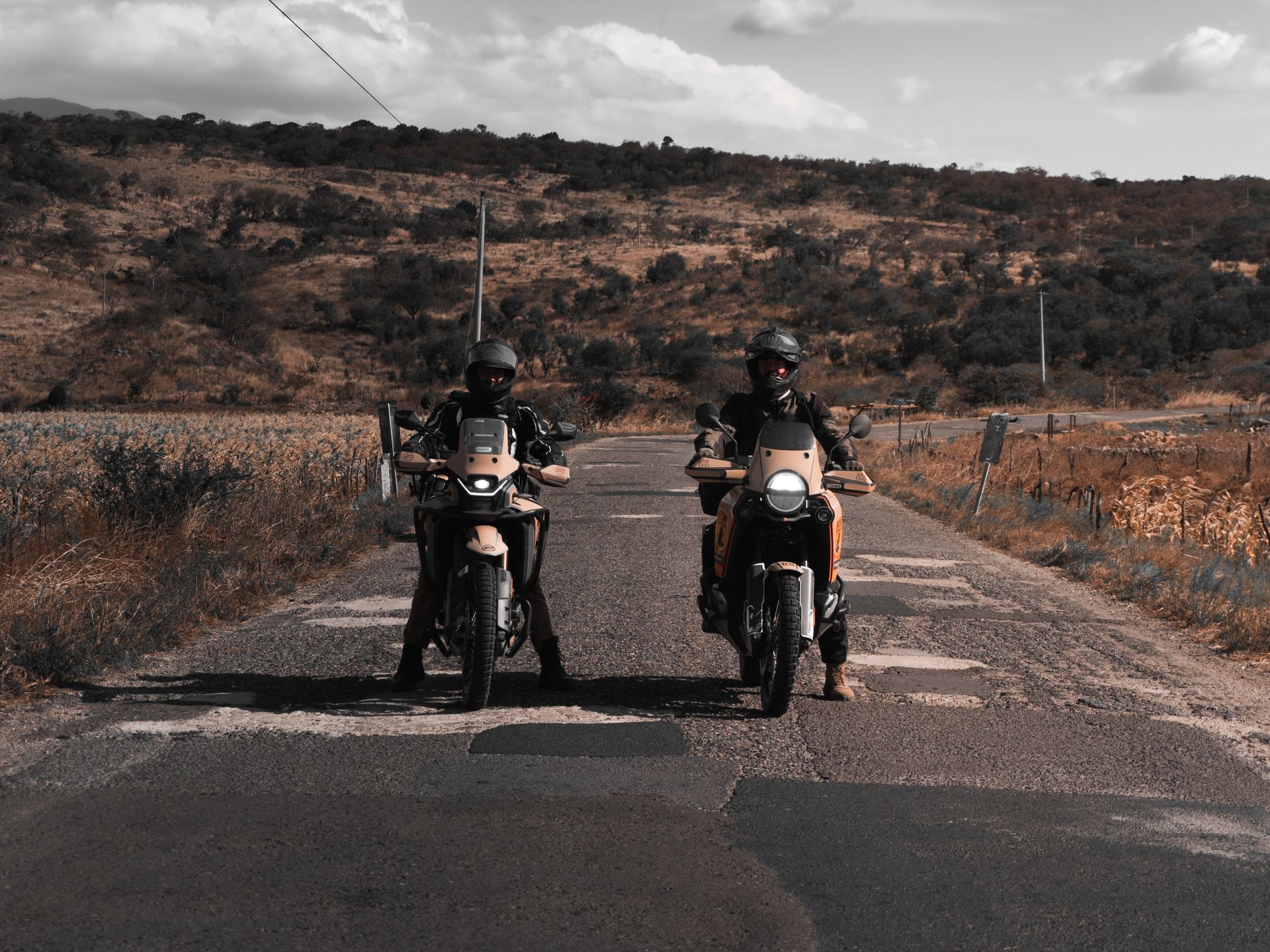 Two motorcyclists on a road with one bike on the shoulder and hills in the background