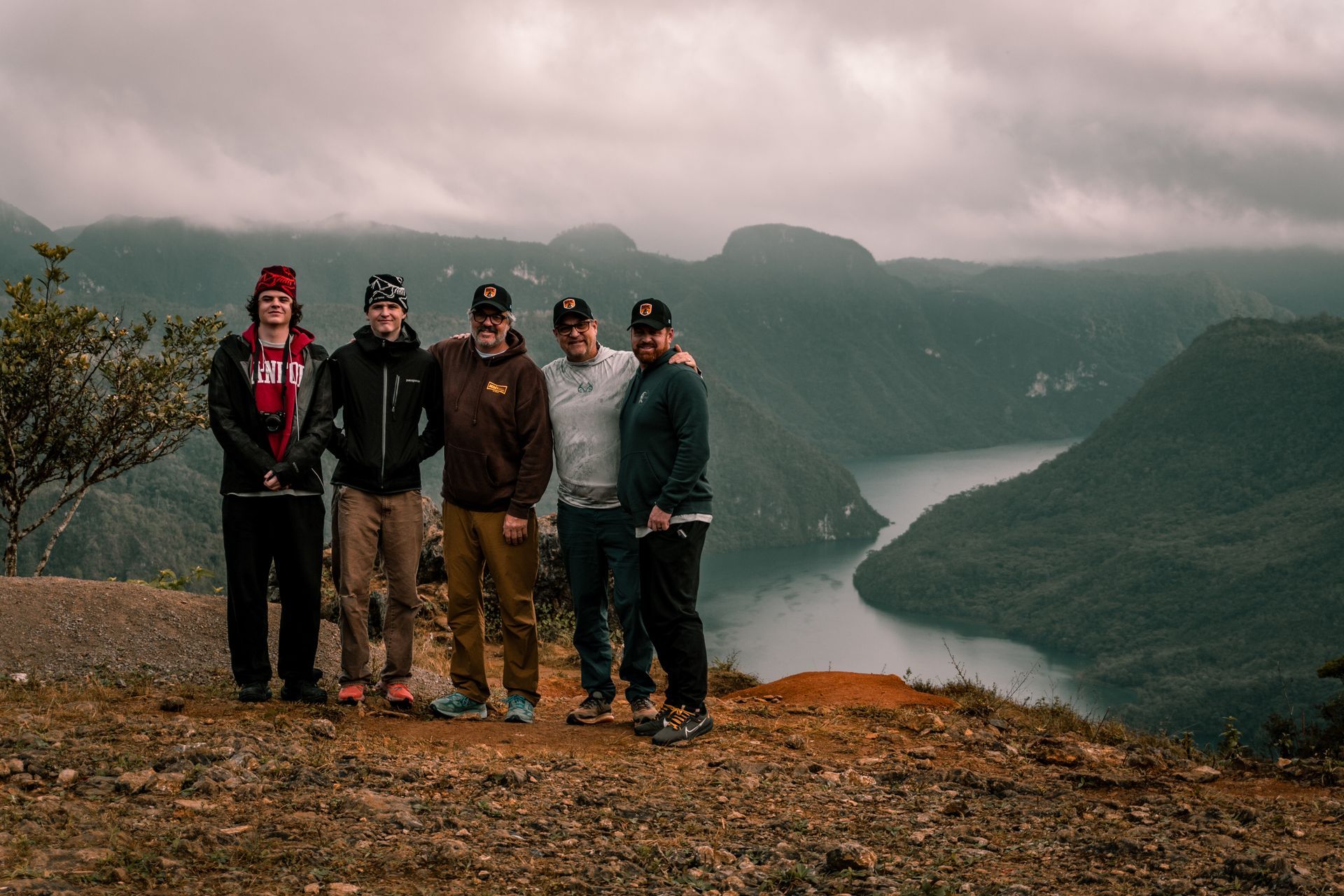 Five people standing on a rocky cliff overlooking a misty mountain lake and forested valley.