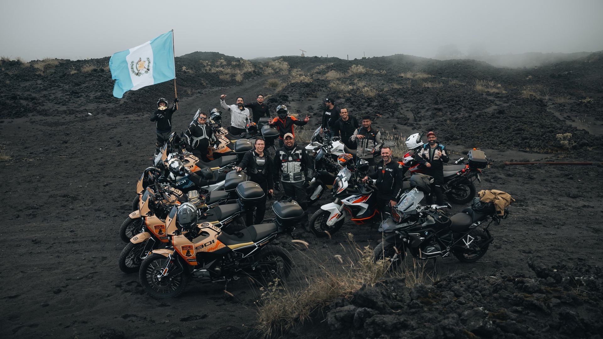 A crowd of motorcyclists gathered on a muddy hillside, with a light blue flag waving above them.