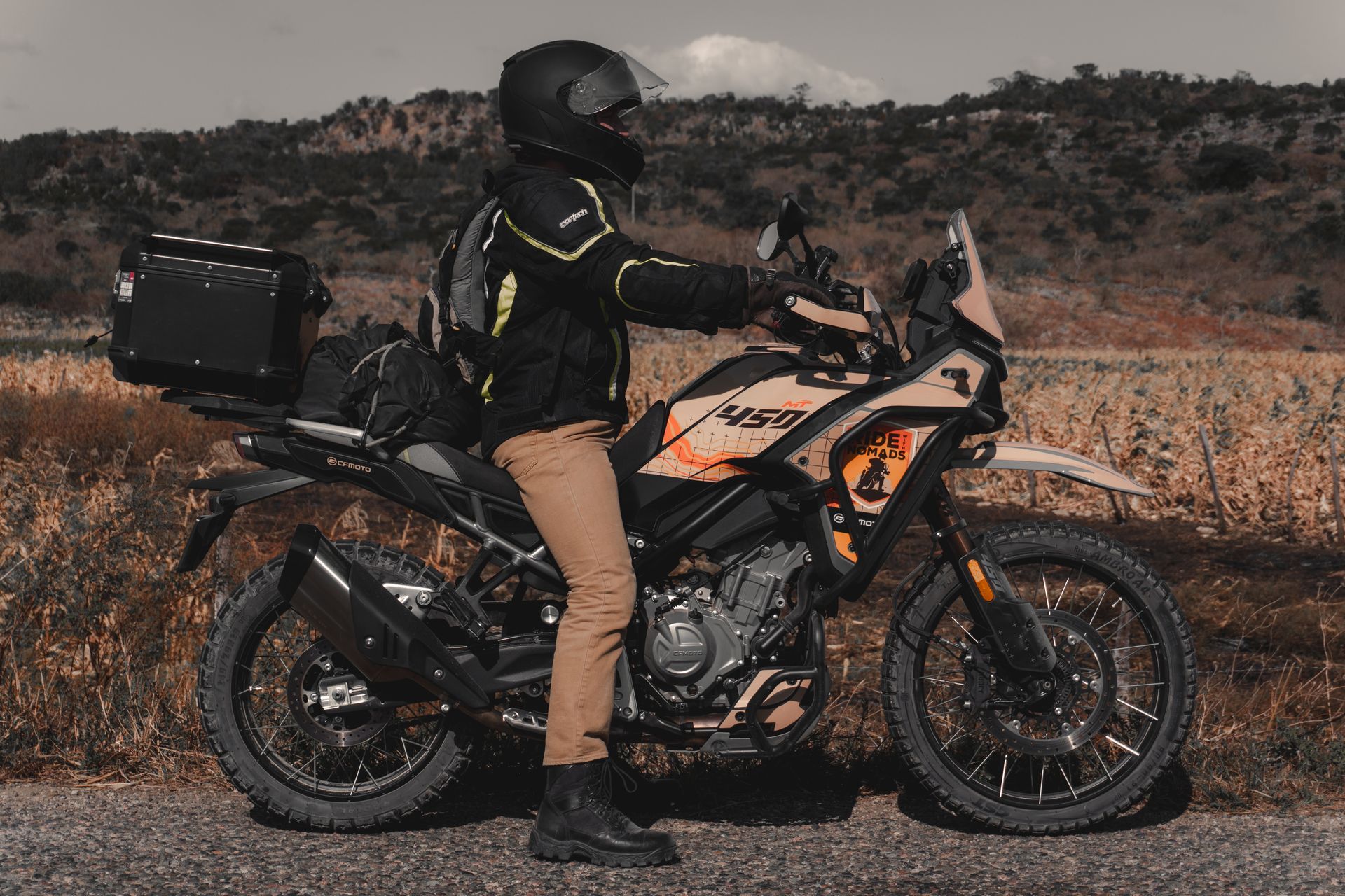 Motorcyclist in black gear on an orange adventure bike parked on a roadside in a dry field