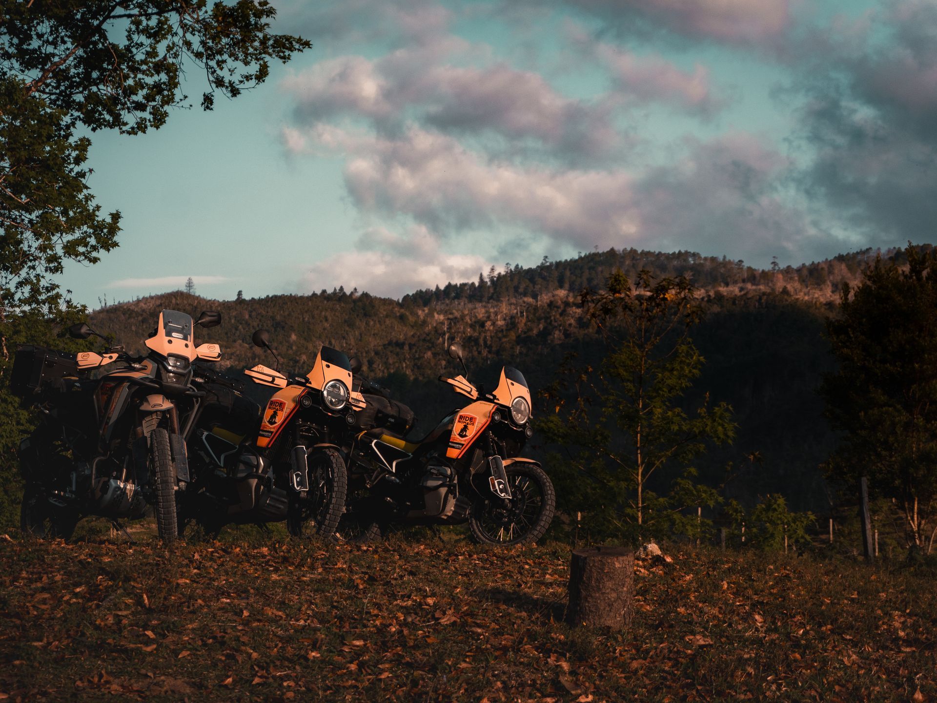 Three dirt bikes parked in a field at sunset with hills and cloudy sky in the background