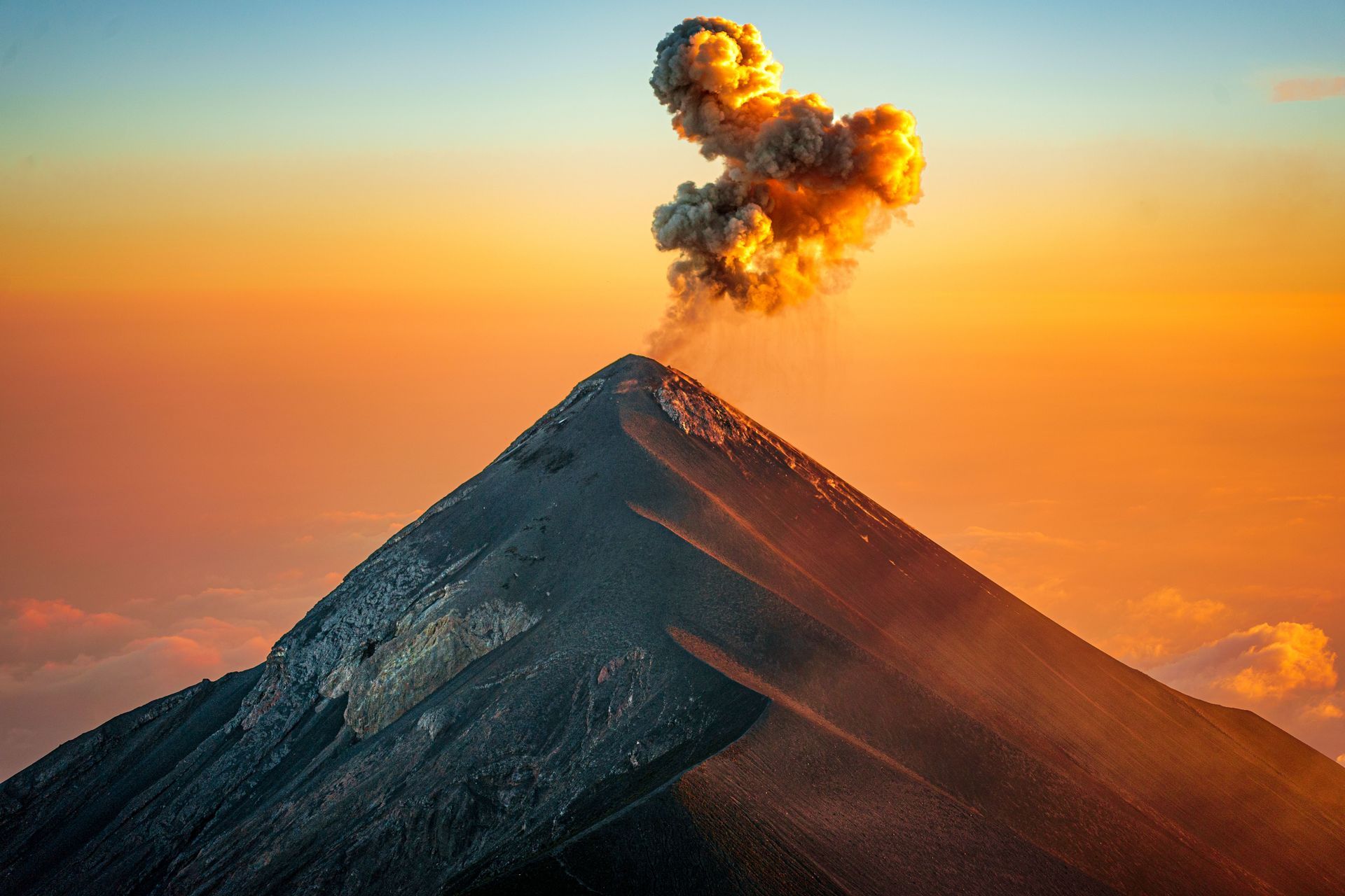 Erupting volcano at sunset with a plume of smoke and glowing orange ash over a dark mountain slope