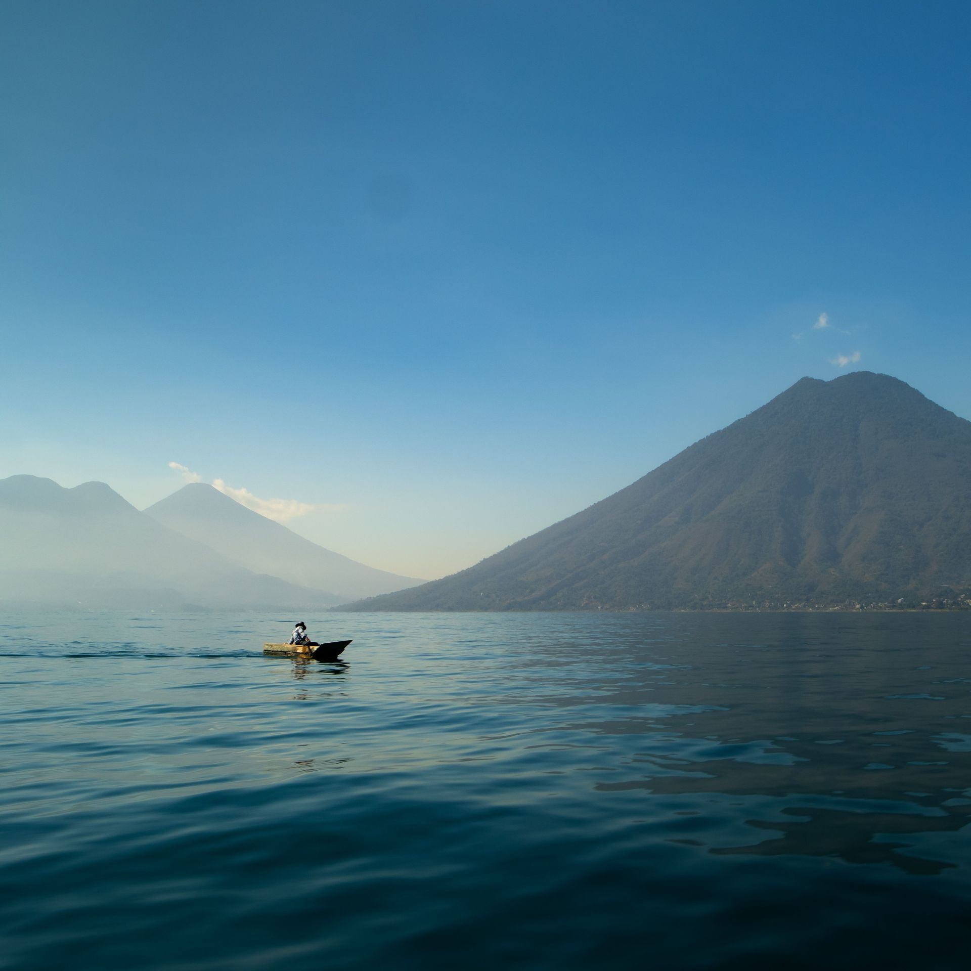 A lone kayaker on calm blue water with misty mountains and a volcano under a clear sky.