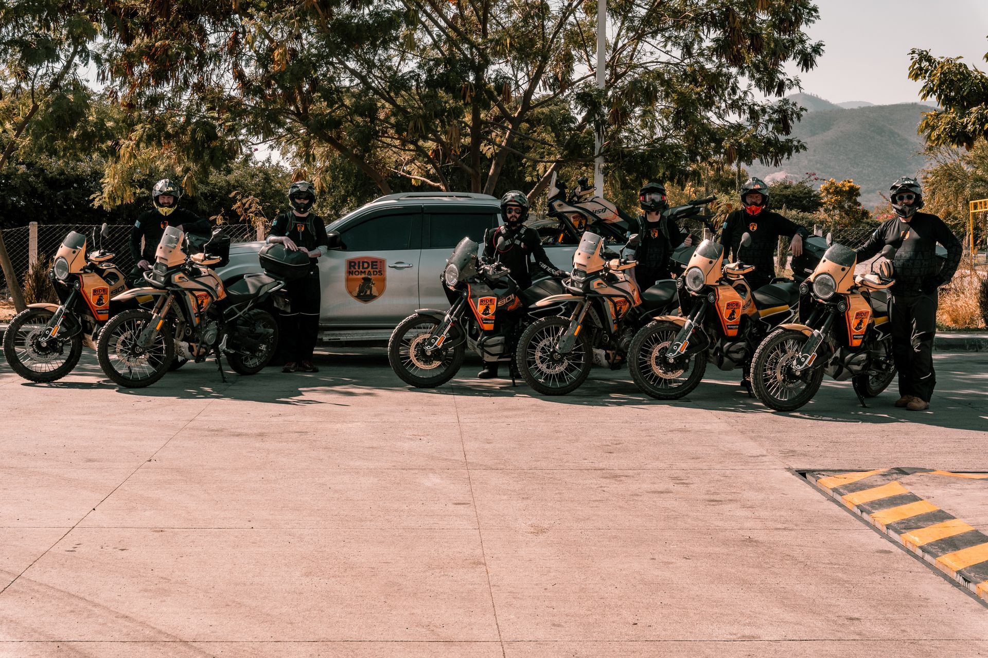 Motorcyclists gathered beside a truck on a roadside, with trees and hills in the background.