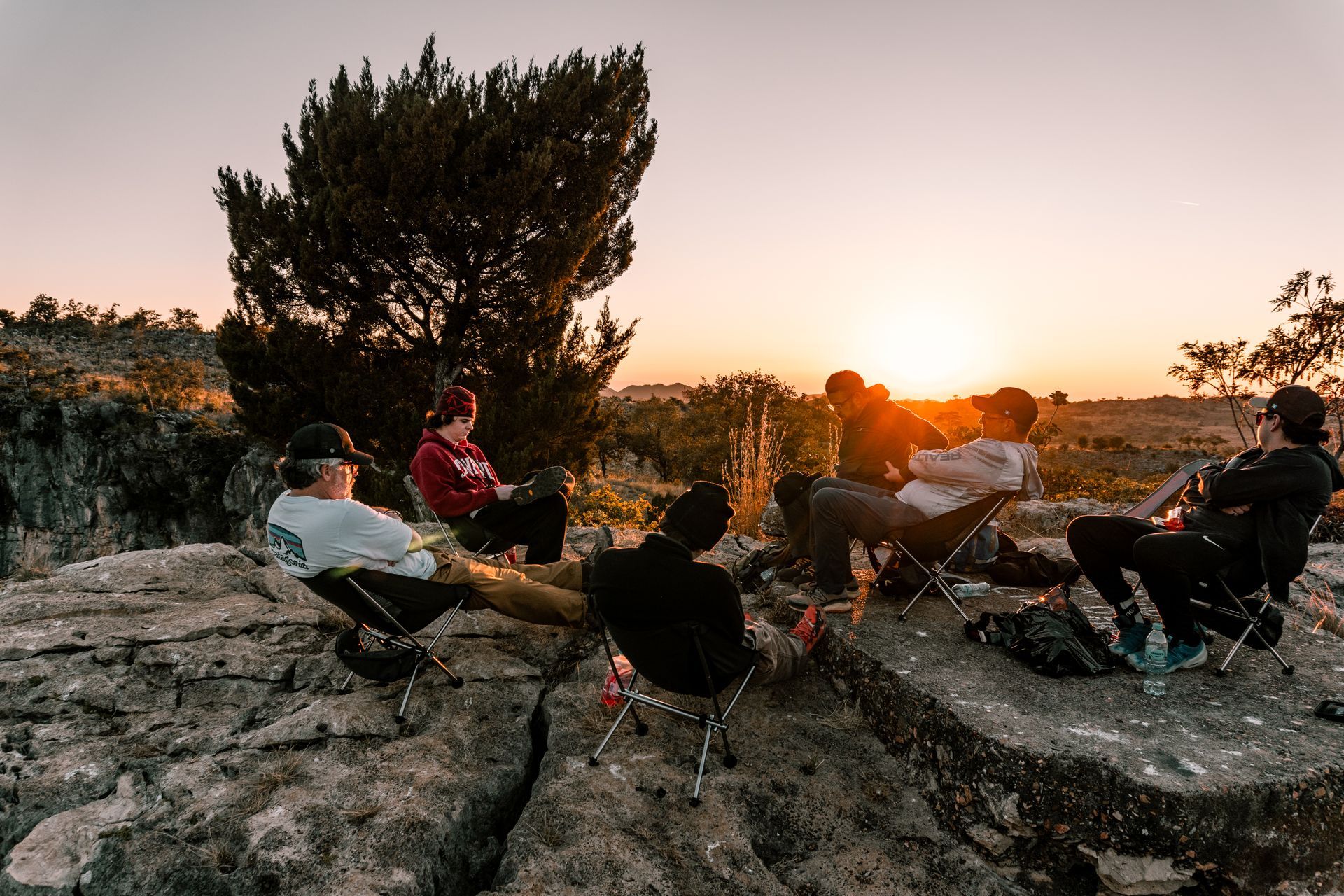 People gathered around a campfire on rocky ground at sunset, with trees silhouetted in the background.