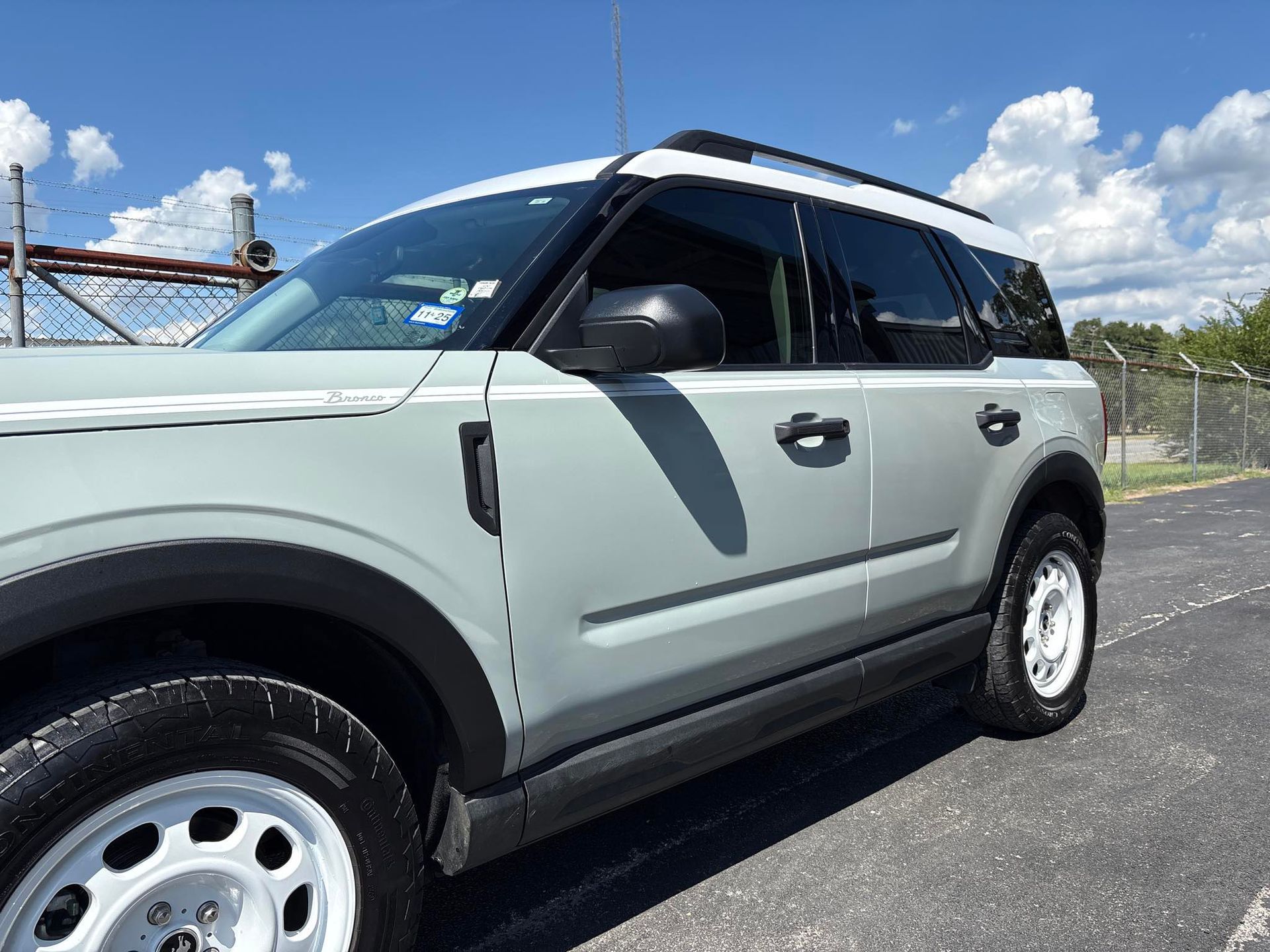 A light gray Ford Bronco Sport parked on asphalt with a white roof and wheels, under a blue sky.