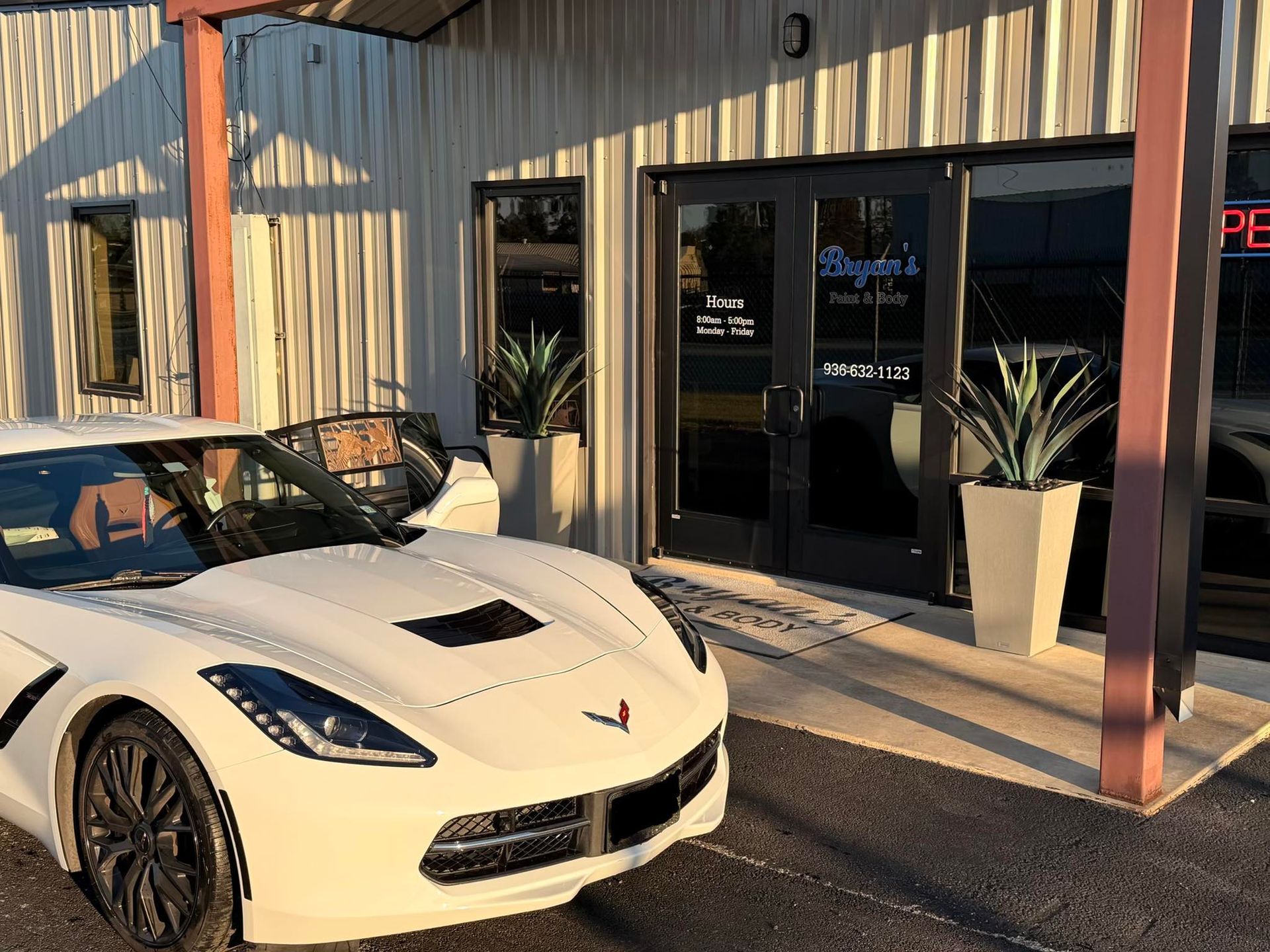 White Corvette parked in front of a restaurant. Double glass doors with text. Potted plants flank the entrance.