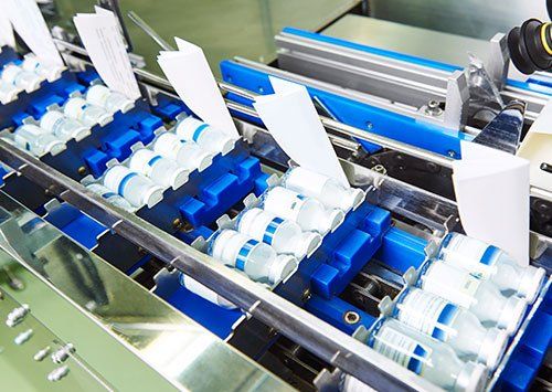 Viales de vacunas en una línea de envasado automatizada de color azul y plata, con etiquetas blancas.