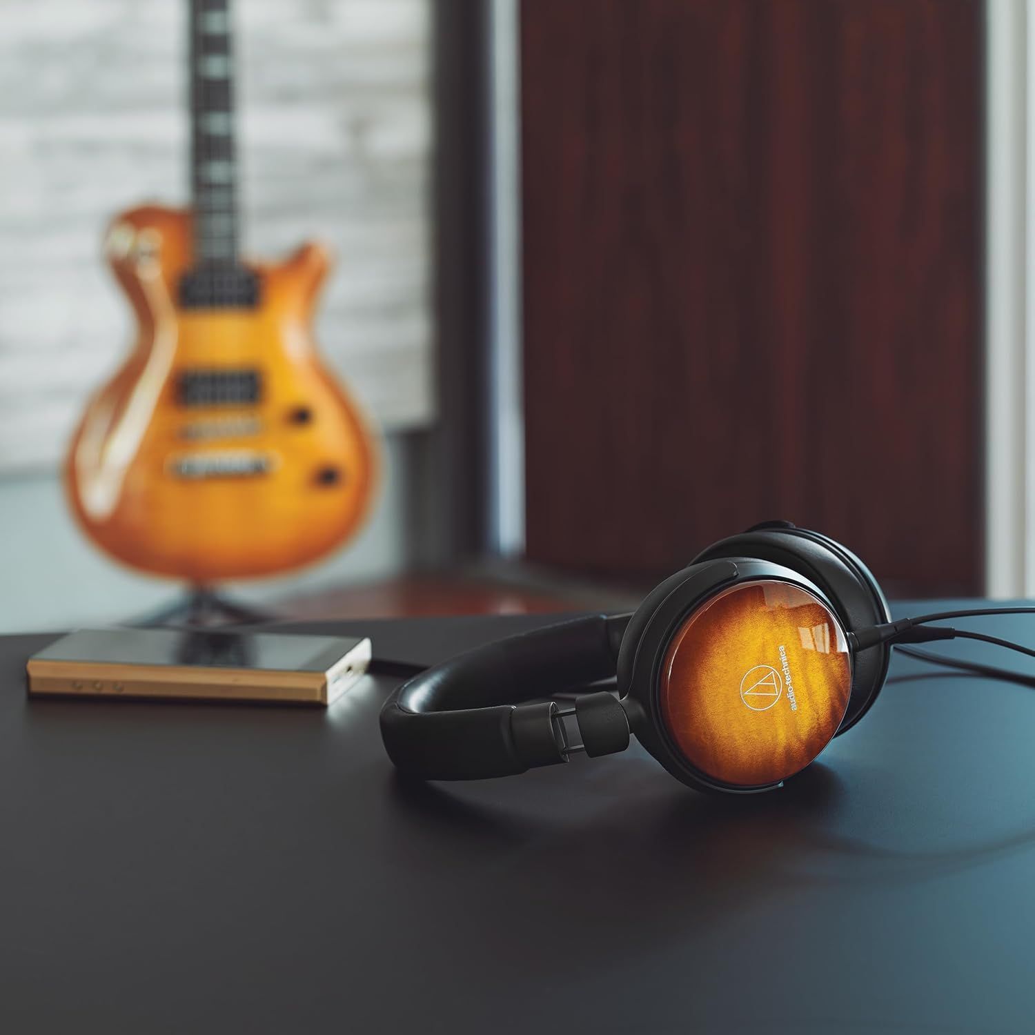 Fone de Ouvido Premium ATH-WP900 Headphones on desk with electric guitar in background.