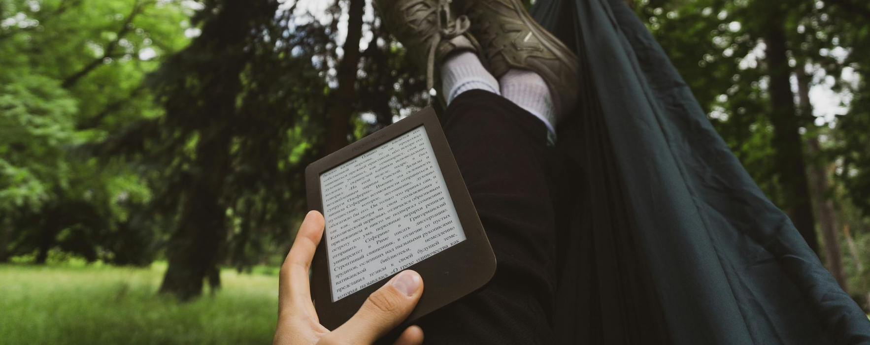 Kindle Unlimited Person holding an e-reader, resting outside in a hammock with trees in the background.