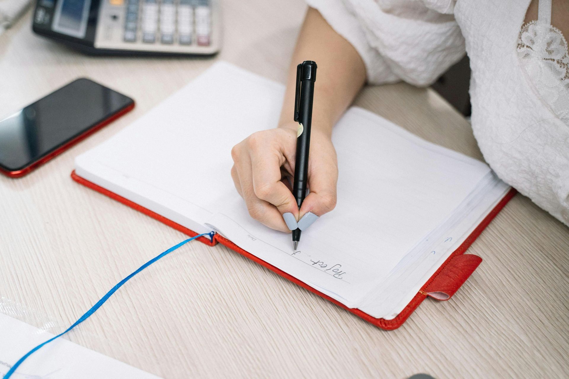 Identificando sua MIT Person writing in a red notebook with a black pen, sitting at a desk. A phone and calculator are visible.
