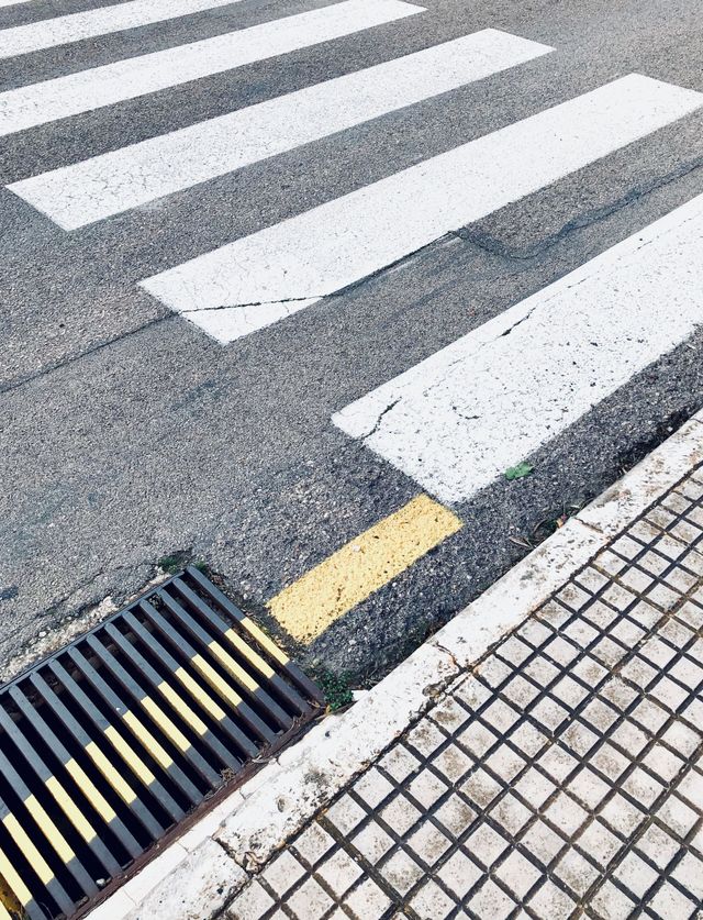 Close-up of a street crosswalk with white stripes, a yellow curb marking, and a storm drain beside tiled pavement.