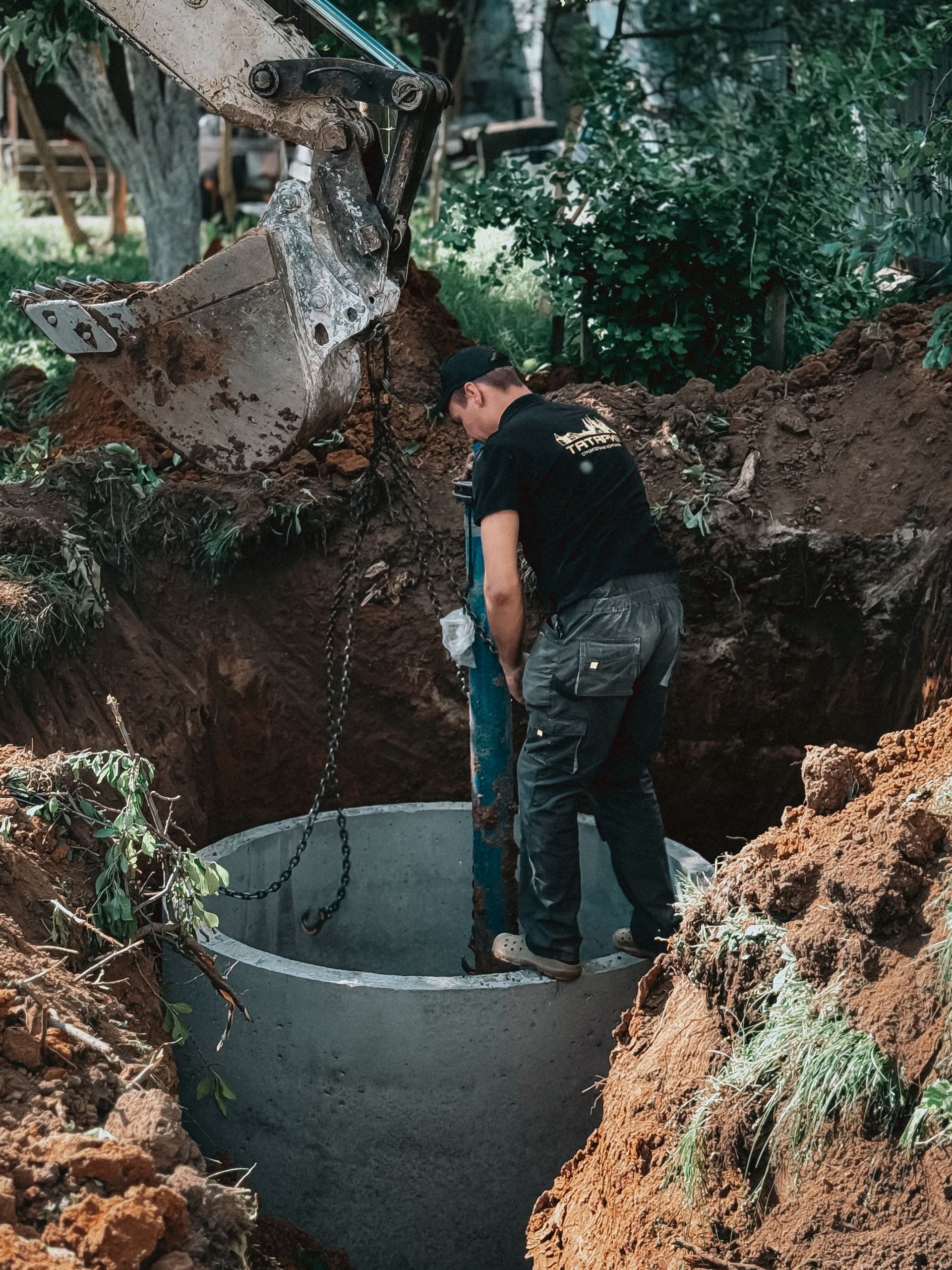 Man in black shirt working inside a deep dirt pit beside a concrete ring outdoors
