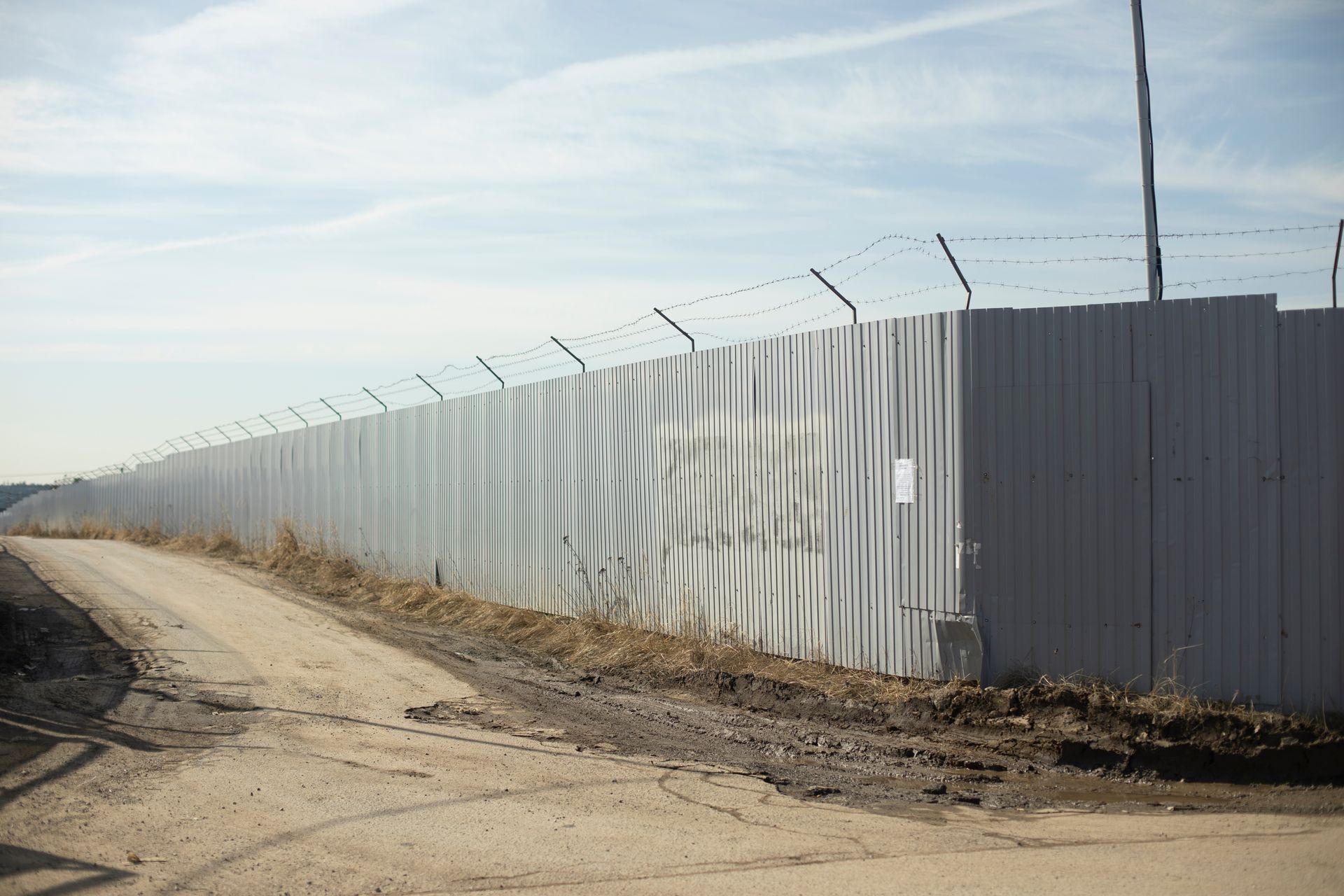Metal privacy fence topped with barbed wire running alongside a dirt road and open land. Metal privacy fence topped with barbed wire running alongside a dirt road and open land.