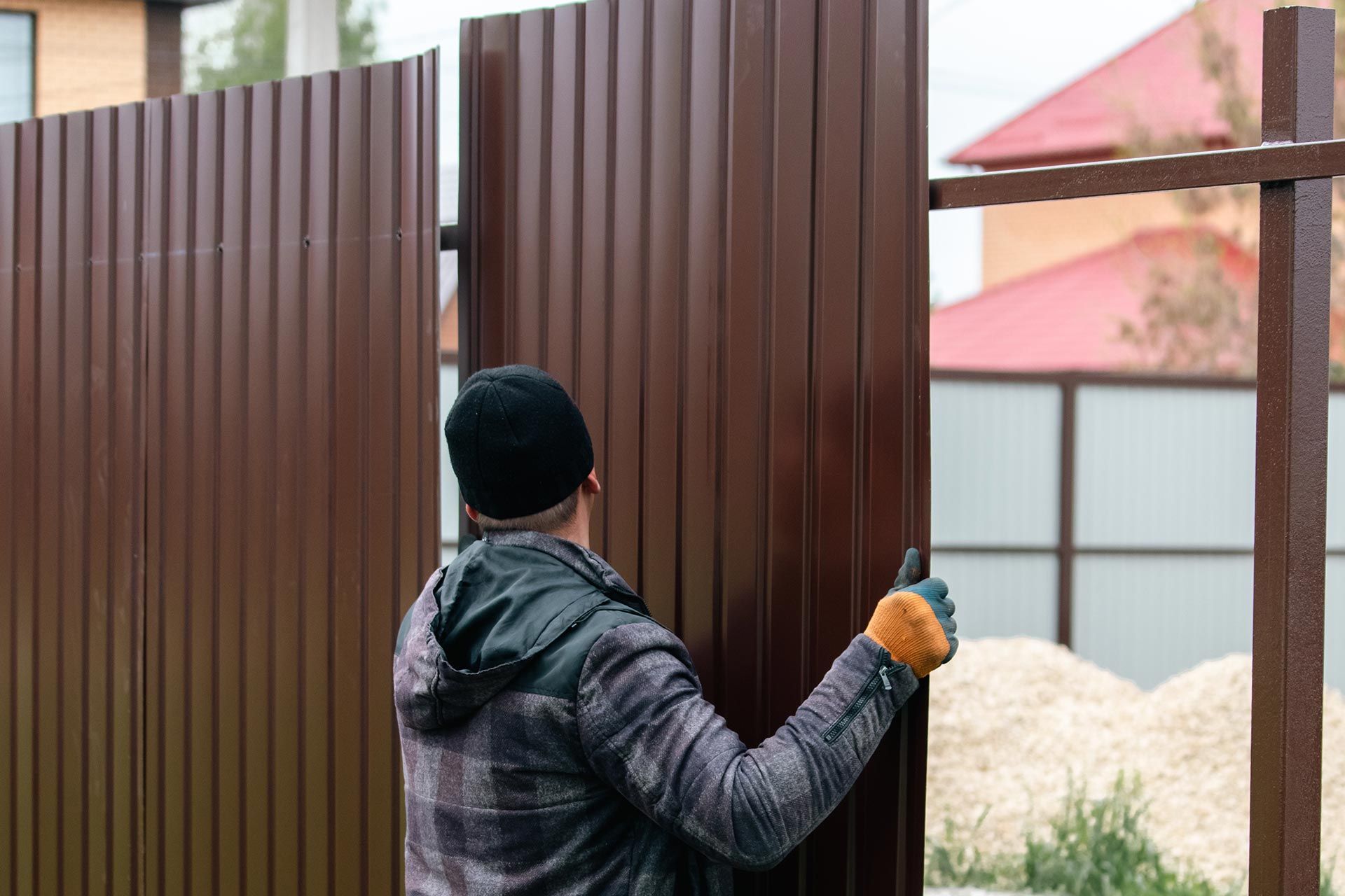 A worker in a plaid jacket installs corrugated brown fence panels in a residential yard.